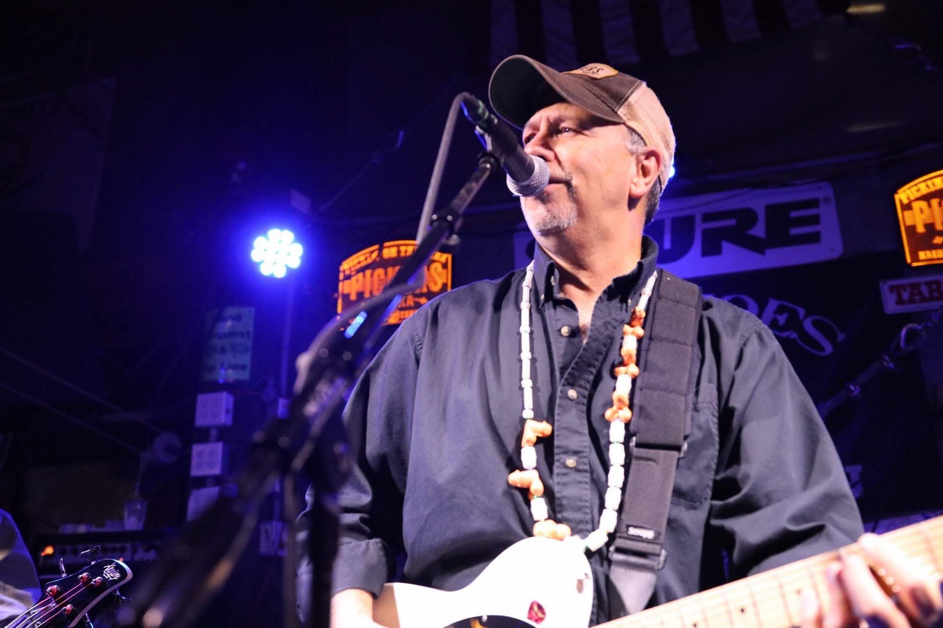 A musician with a baseball cap singing into a microphone while playing an electric guitar on a dimly lit concert stage.
