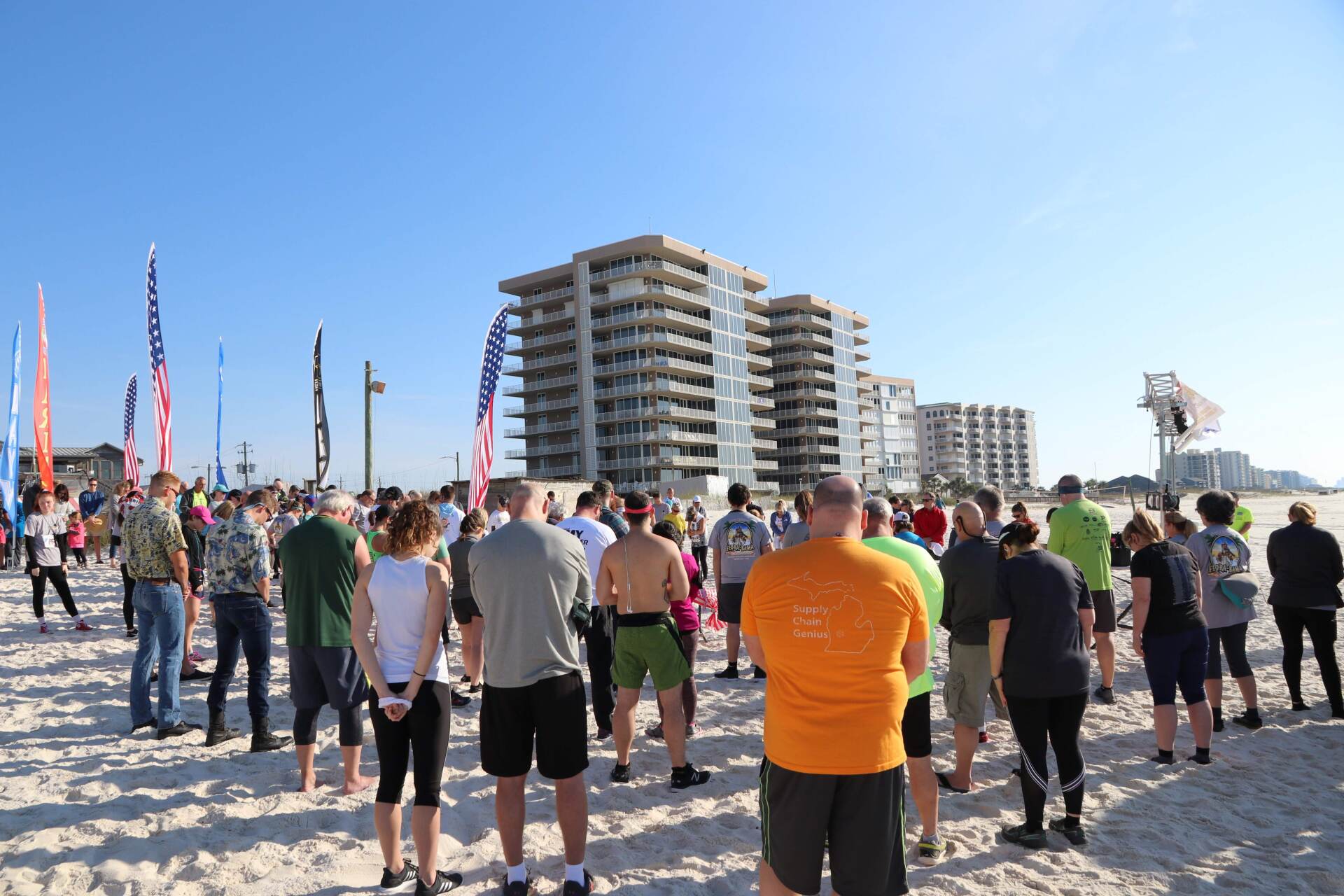 A crowd stands on a beach facing high-rise buildings, with several flags displayed in the sand.