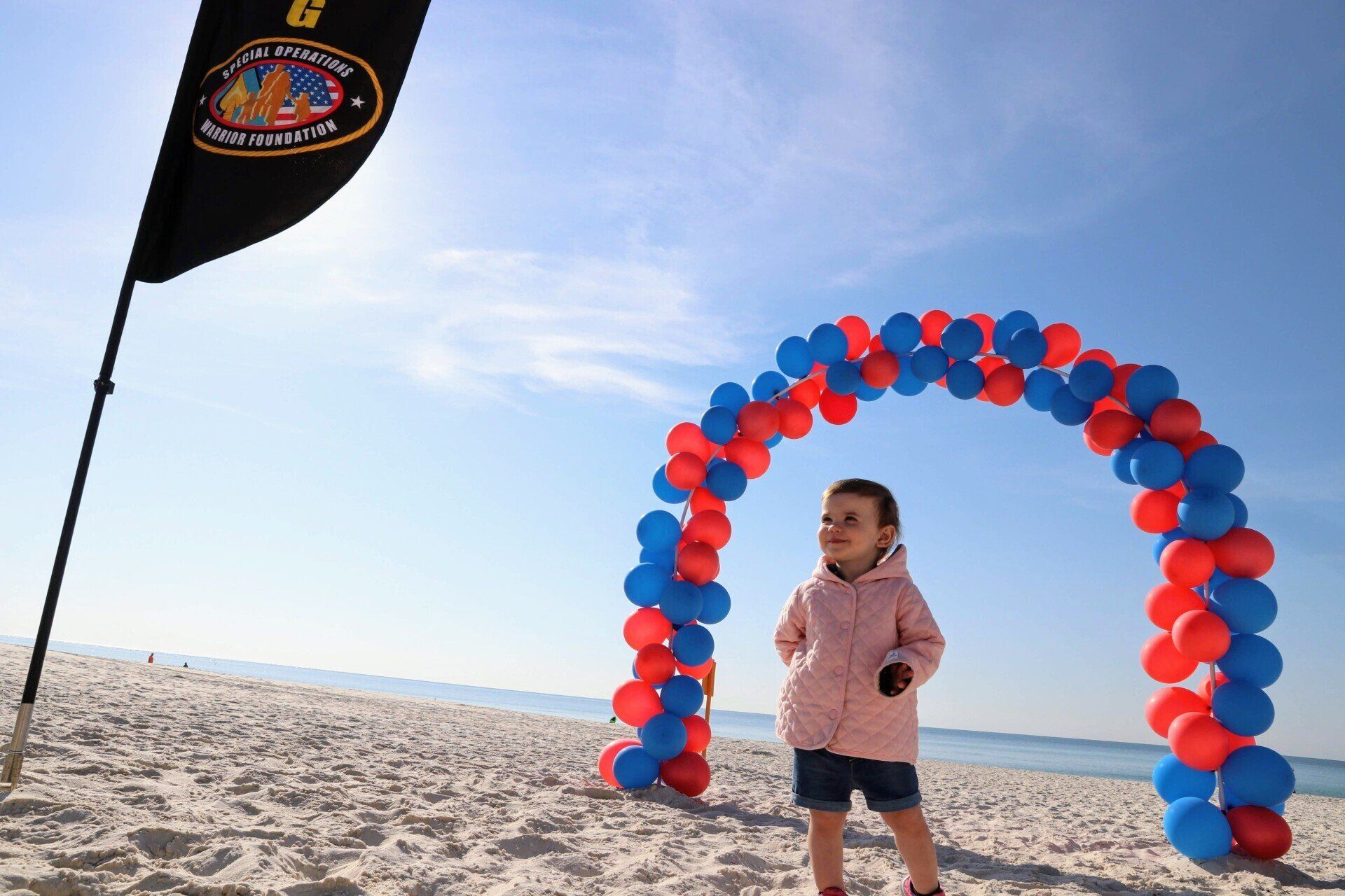 A child stands under a red and blue balloon arch on a sandy beach near a black flag.