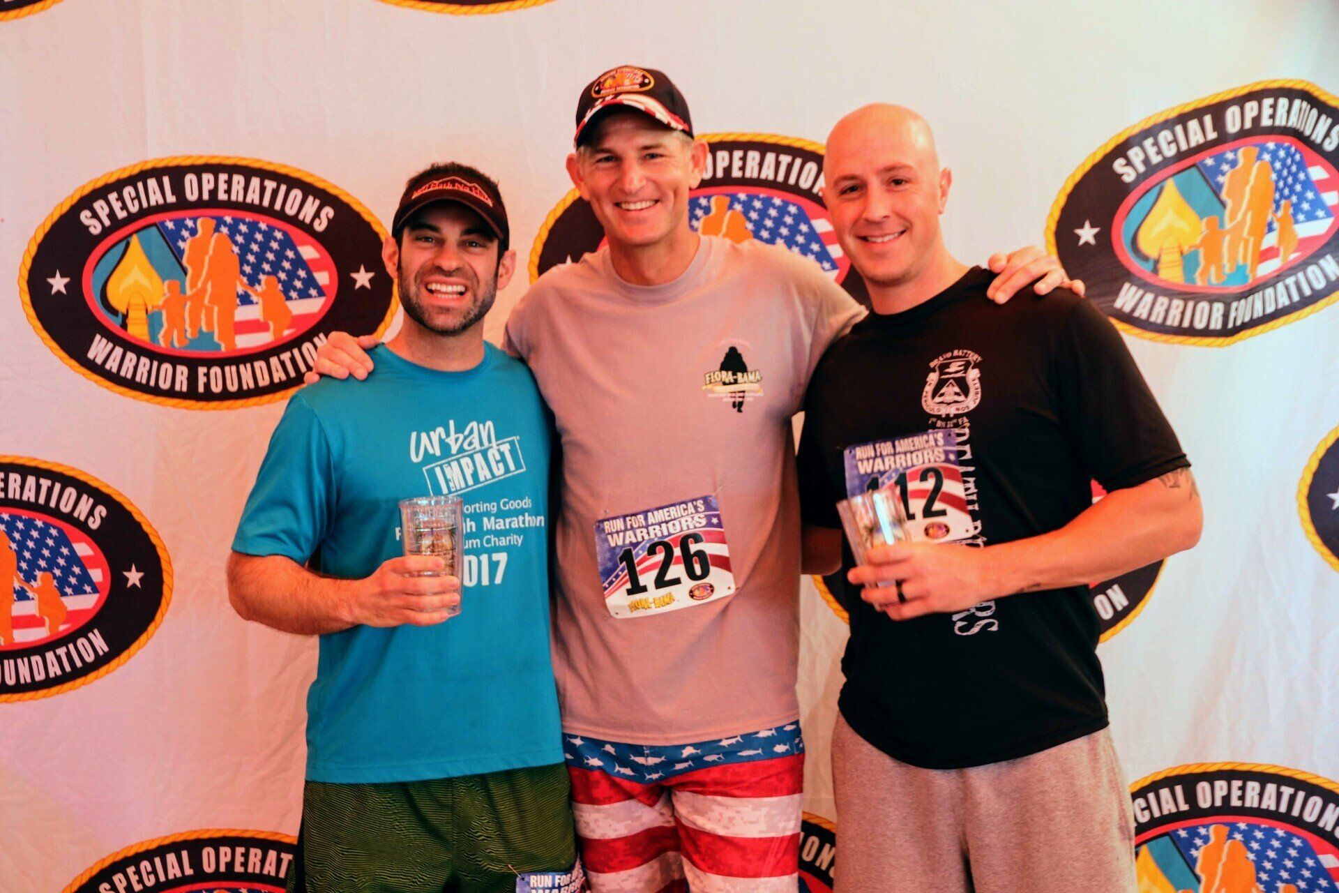 Three men pose smiling in front of a Special Operations Warrior Foundation banner, holding small cups.
