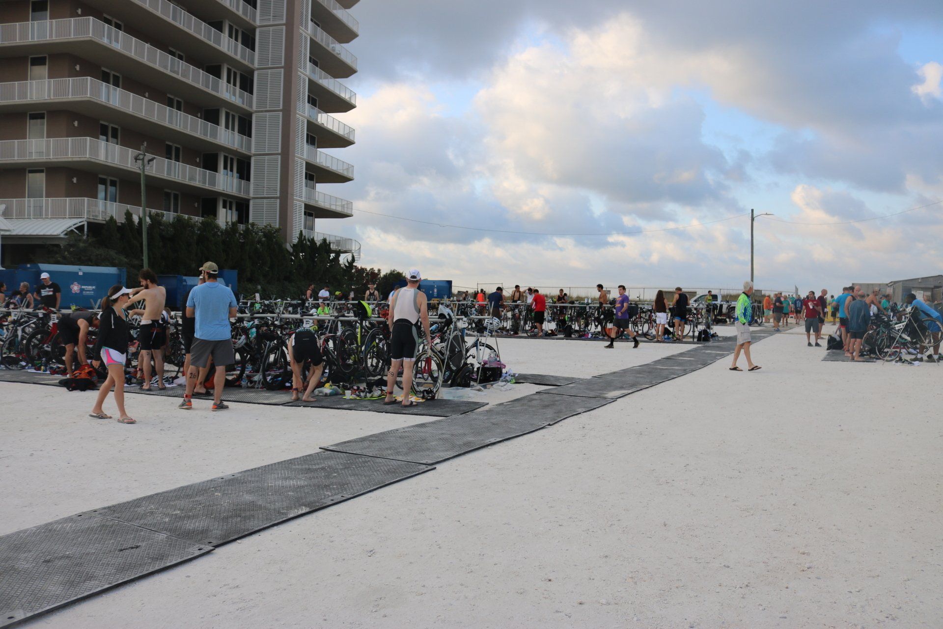 Athletes set up bicycles in a transition area on a sandy beach near a large apartment building during a race event.