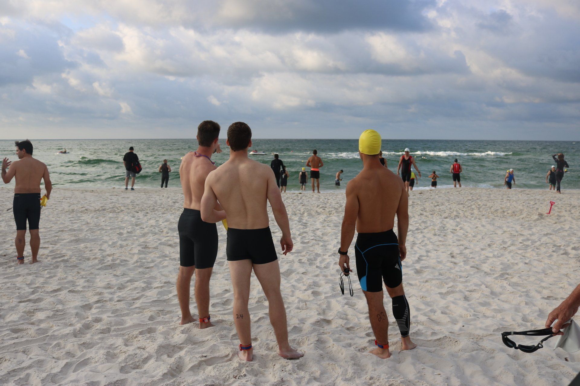 Several people in swimwear stand on a white sand beach looking out at the ocean under a cloudy sky.