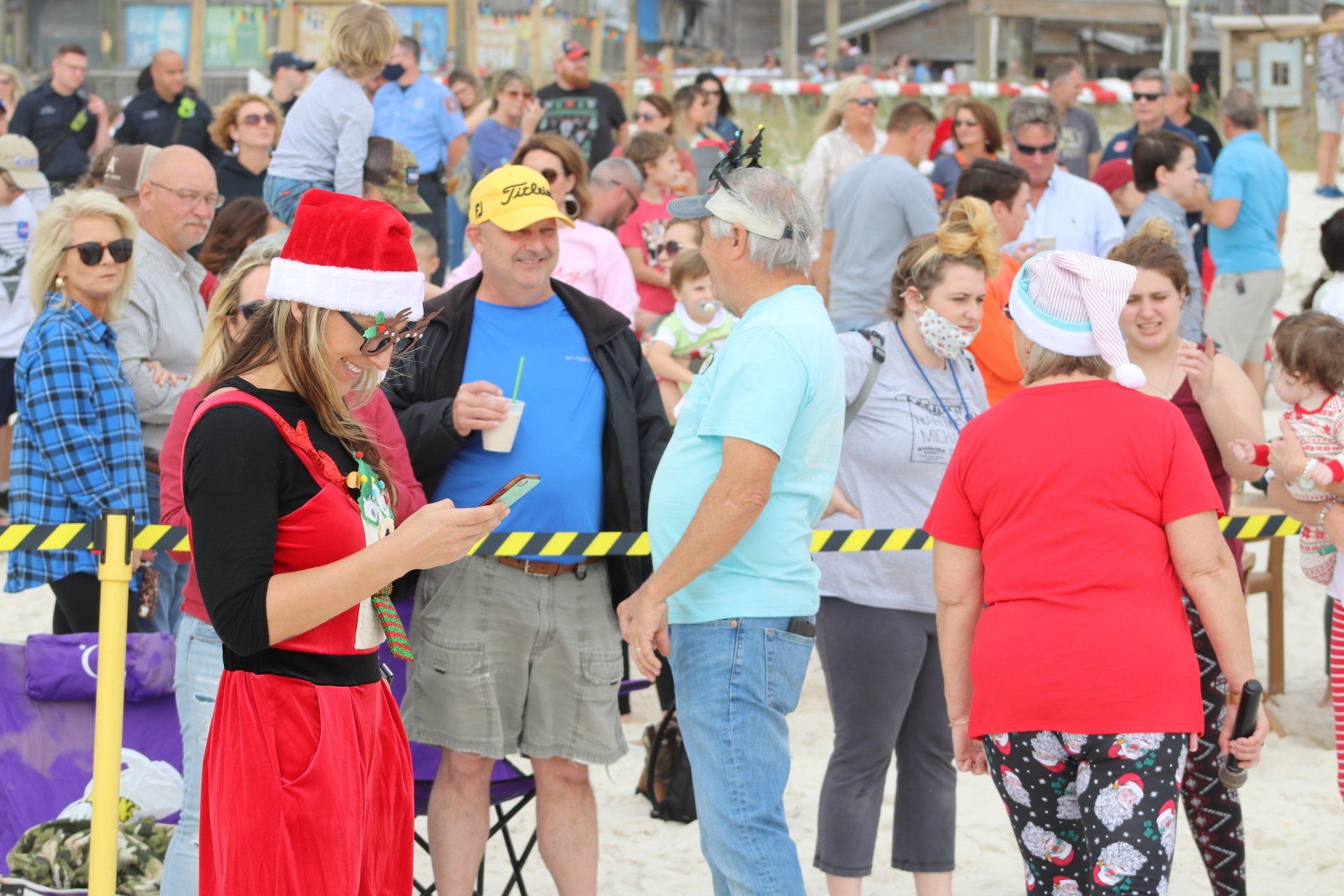 A crowd gathers on a sandy beach for a holiday event, with many attendees wearing Santa hats or festive clothing.