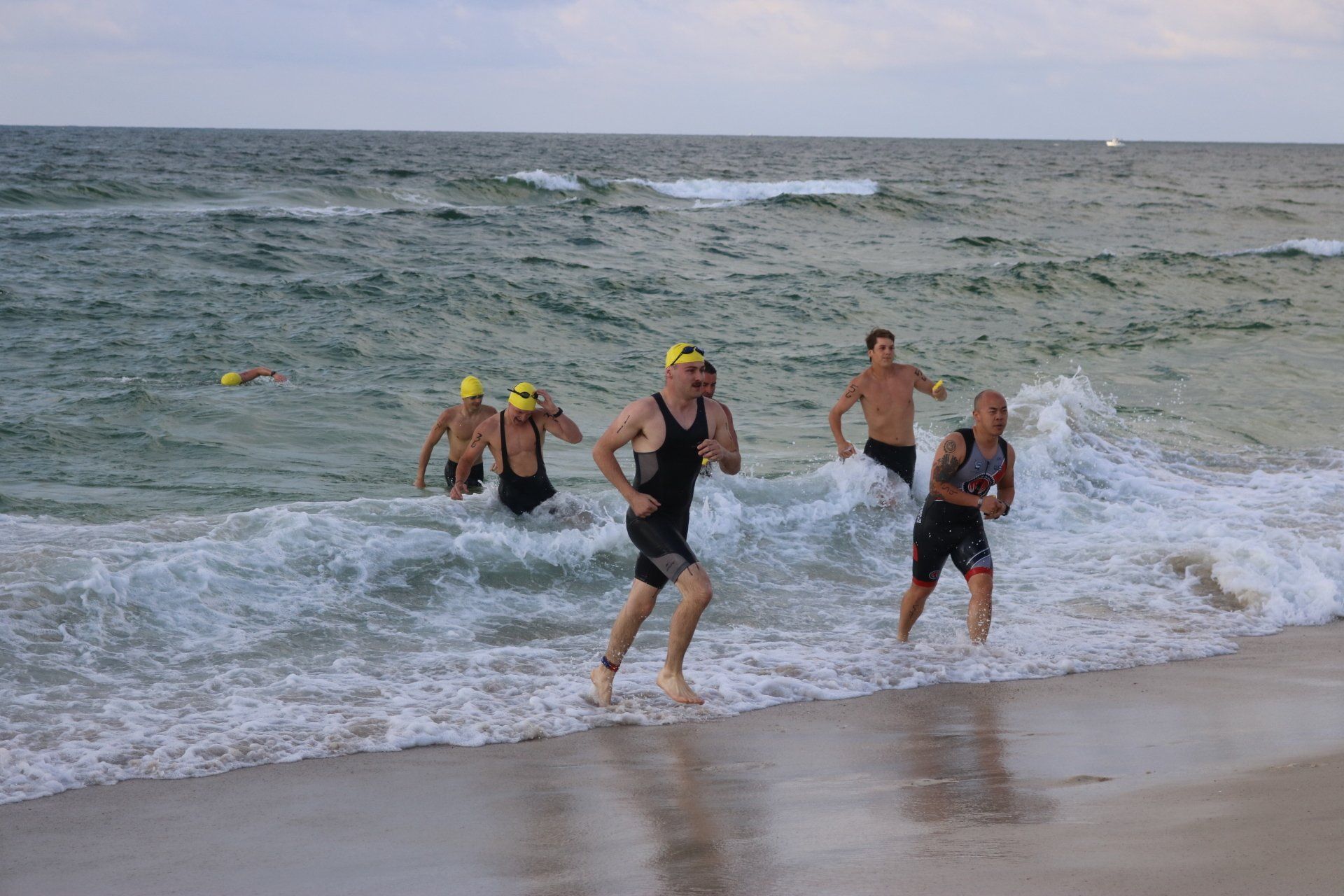 Participants in swim gear run out of the surf and onto a sandy beach after an ocean swim.