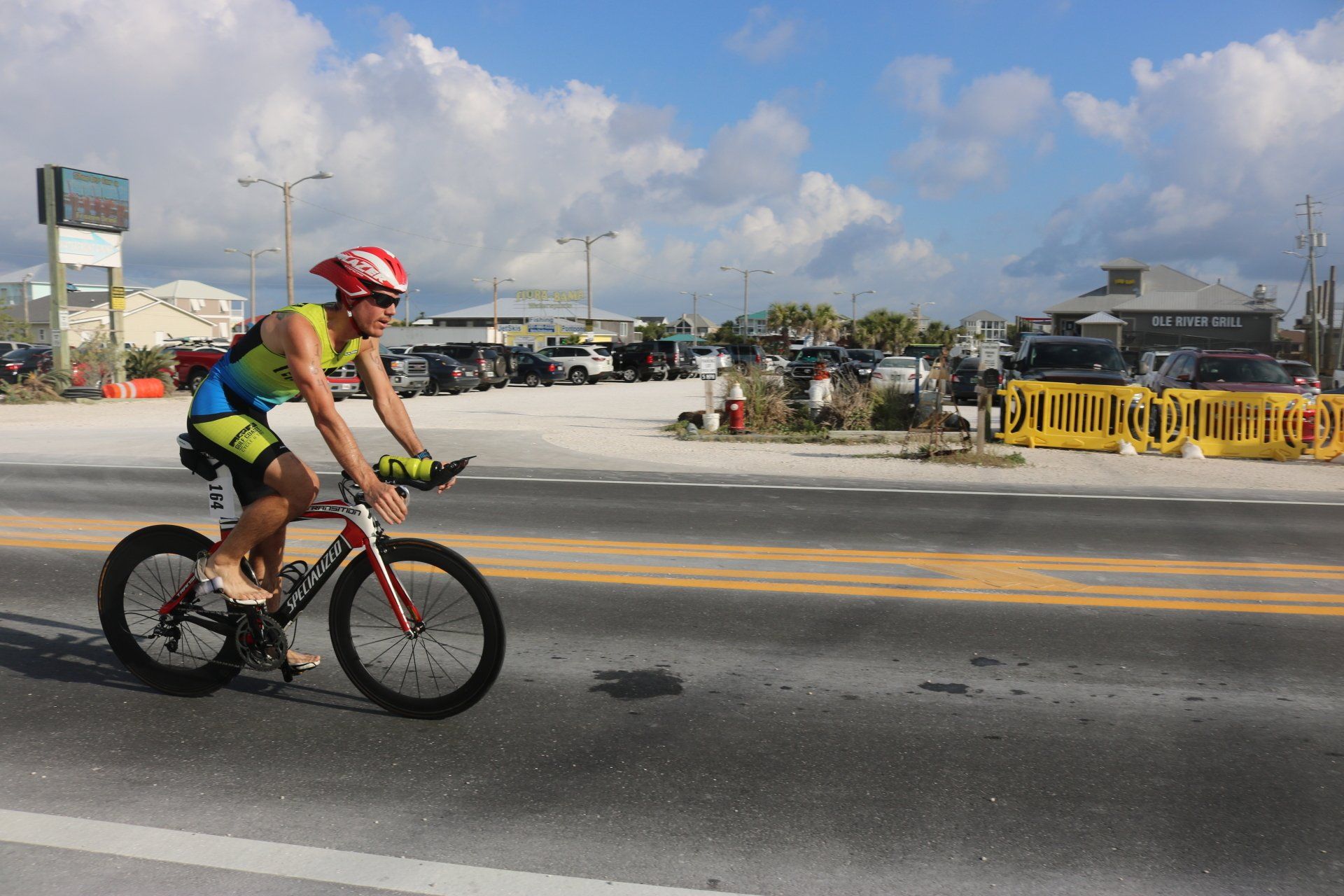A cyclist in a colorful athletic suit and red helmet rides along a road near a parking lot under a cloudy sky.