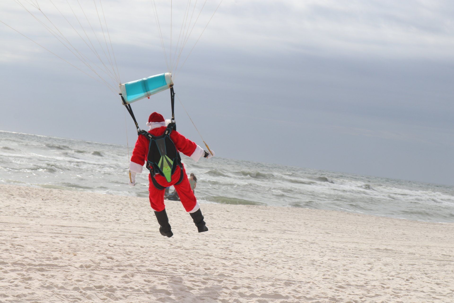 A person dressed as Santa Claus kitesurfing on a sandy beach near the ocean.
