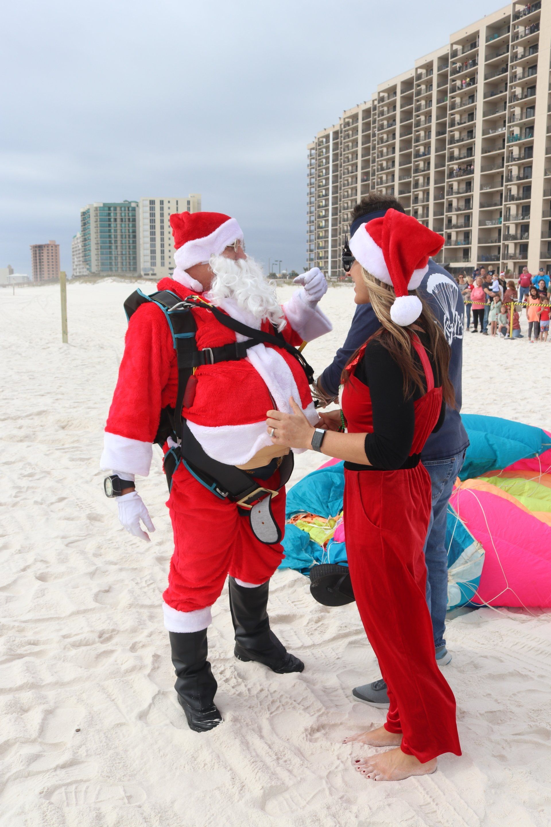 Santa and a person in holiday attire standing on a sandy beach next to a colorful parachute after landing.