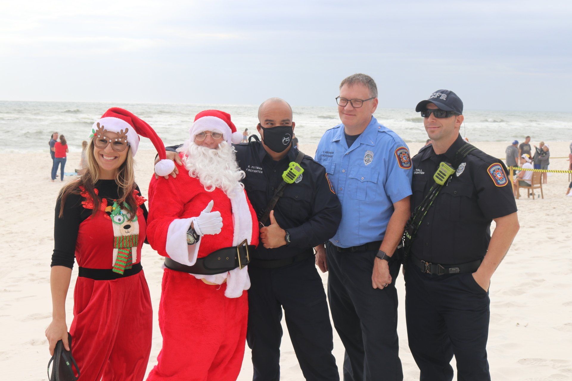A woman in a holiday outfit, Santa Claus, and three law enforcement officers pose for a photo on a sandy beach.