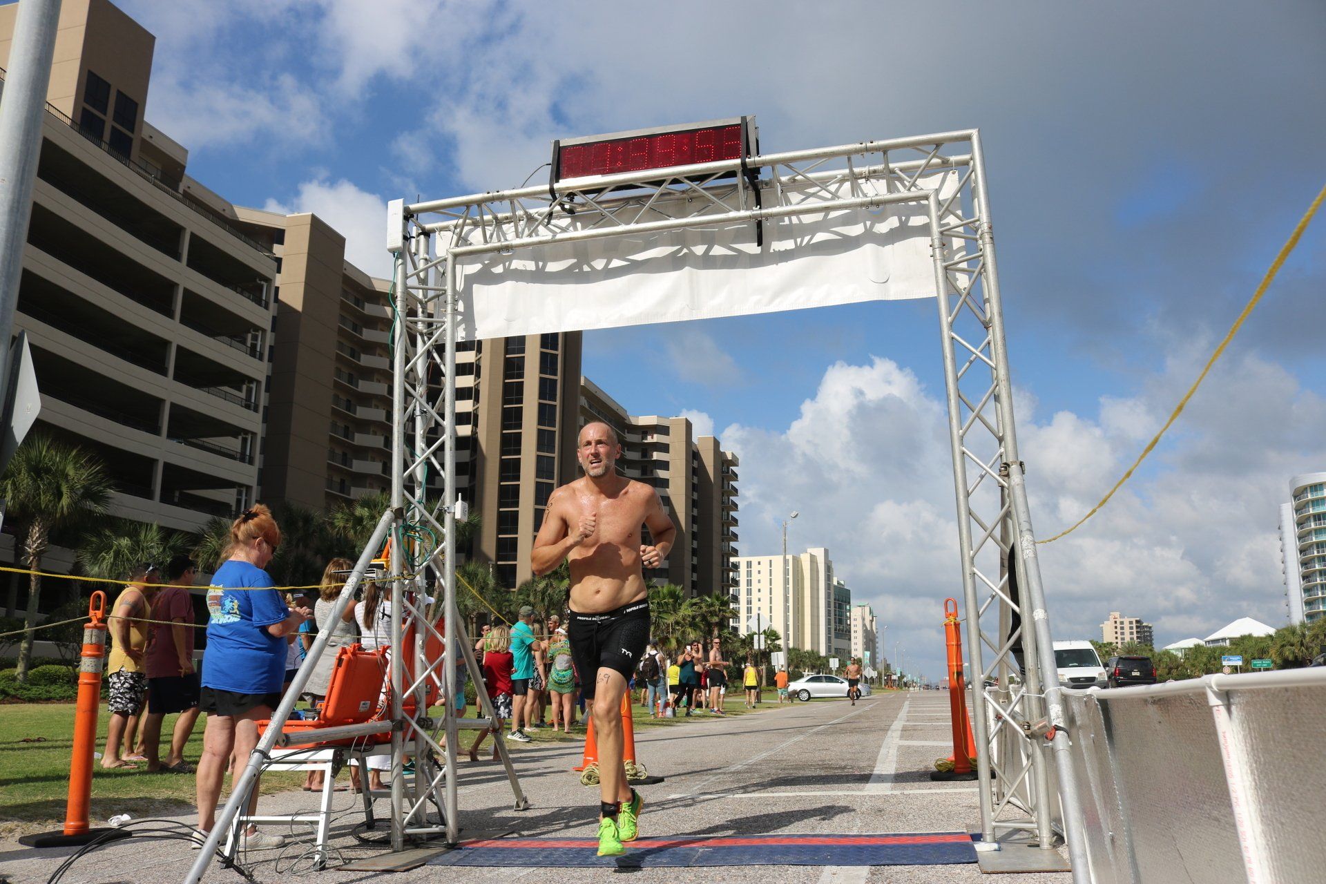 A shirtless runner crosses a finish line on a road near beachfront buildings under a bright, partly cloudy sky.