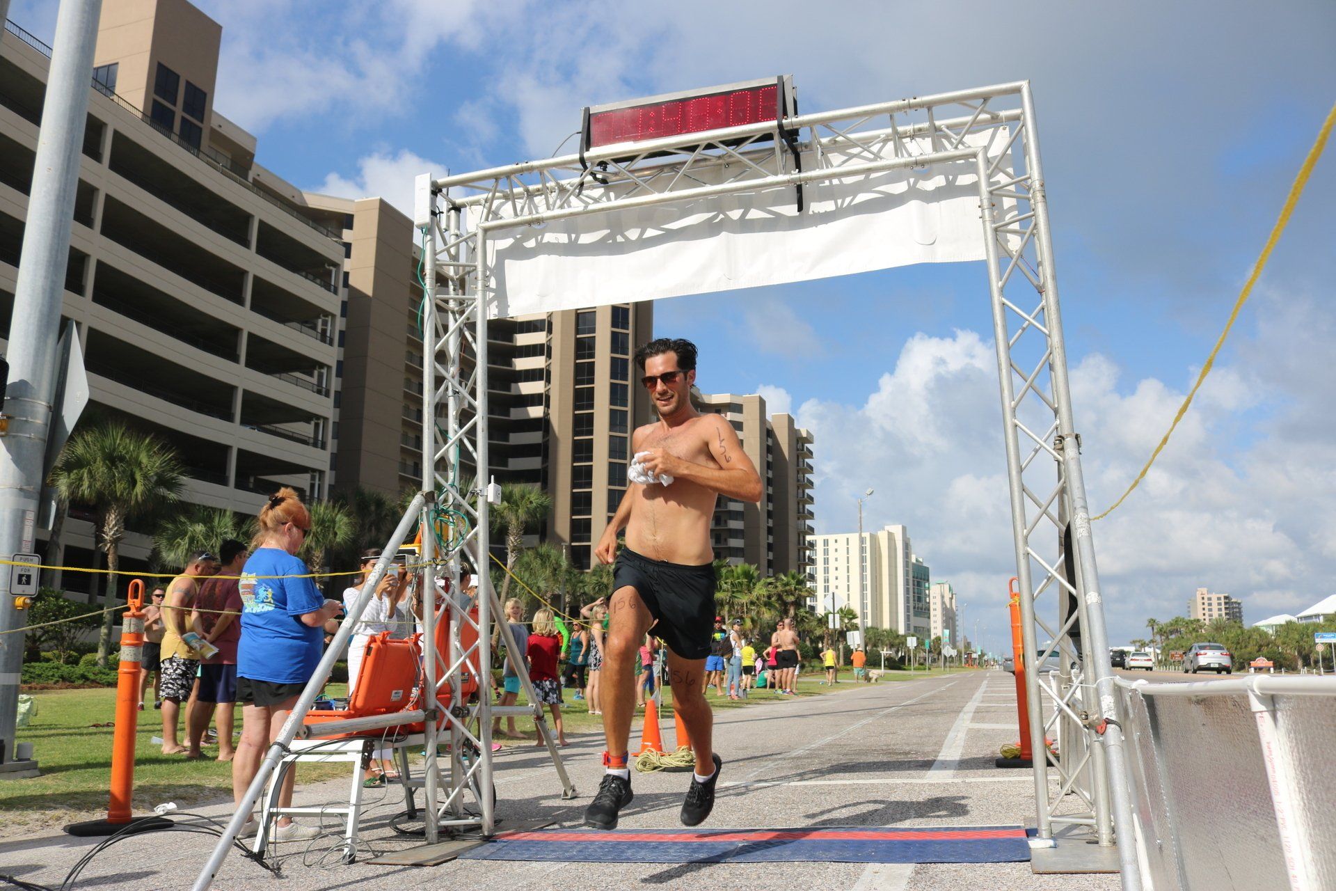 A person runs shirtless through a finish line arch on a road near a beachside hotel.