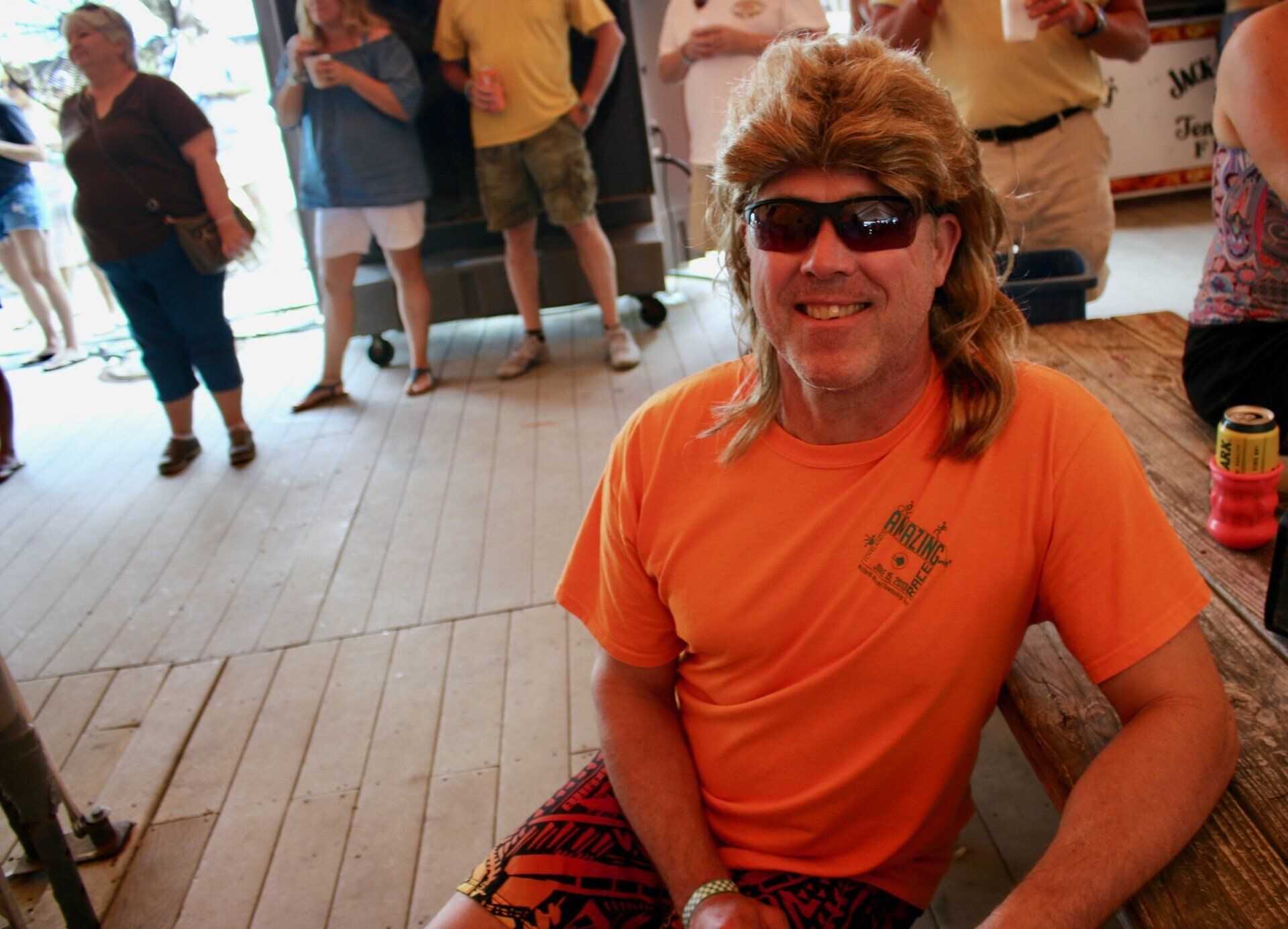 A smiling person with a mullet haircut wearing orange sunglasses and a shirt sits at a table near a group of people.