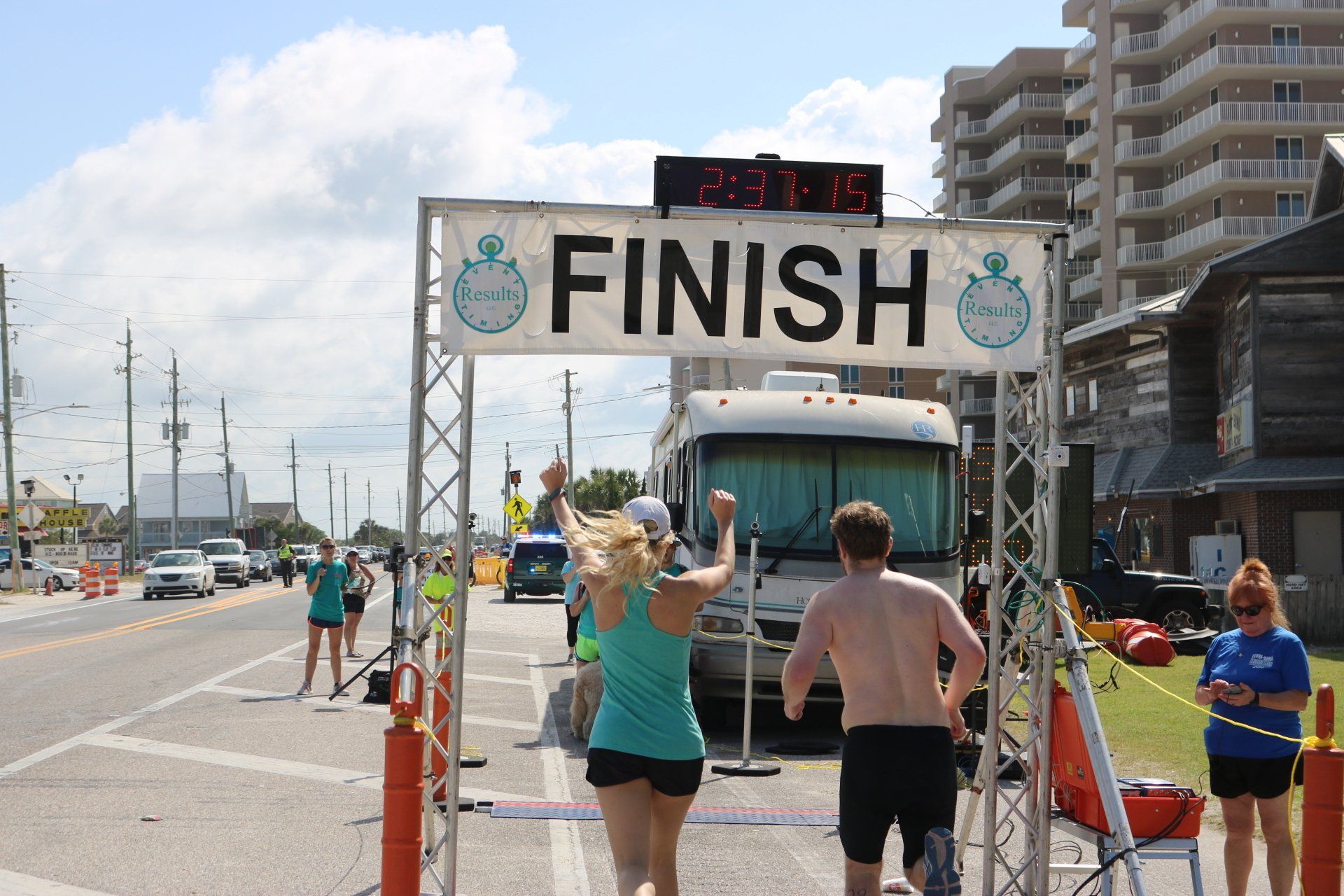 A finish line arch over a road with runners crossing it under a bright sky, next to a large parked recreational vehicle.