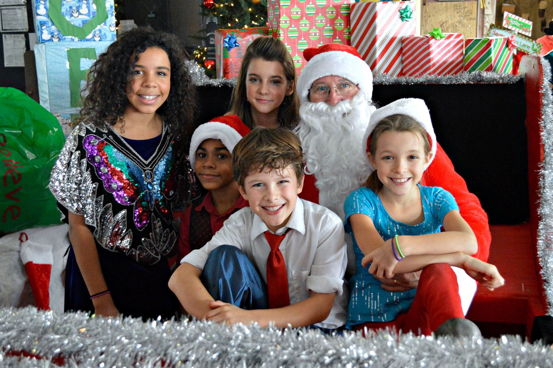 A group of children posing for a photo with Santa Claus, surrounded by festive Christmas decorations and wrapped gifts.