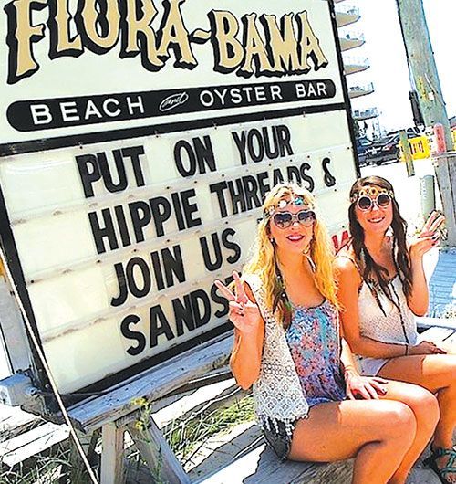 Two people dressed in hippie-style clothing pose in front of a Flora-Bama Beach Oyster Bar sign advertising an event.