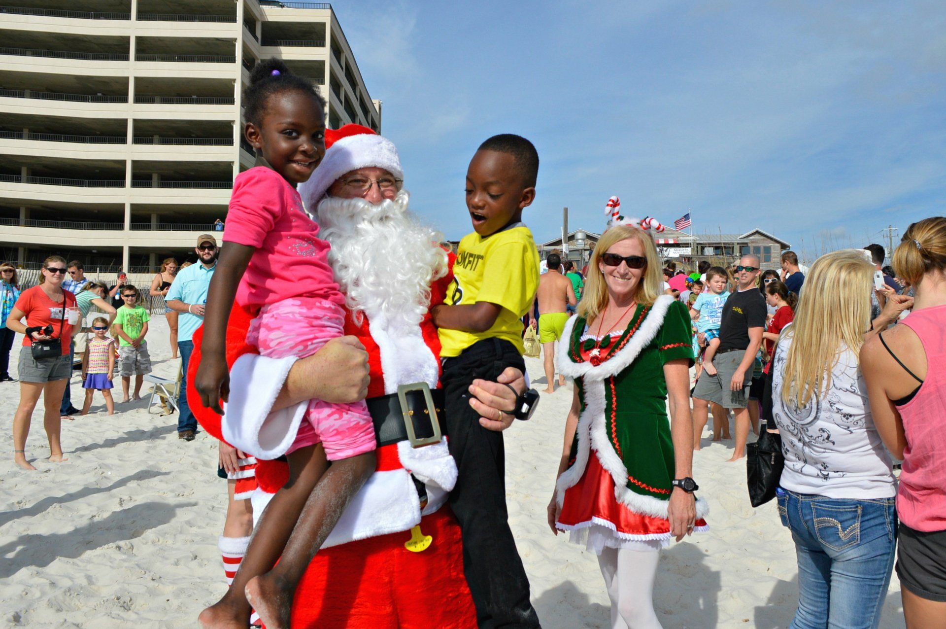 Santa Claus and a festive helper on a beach, holding two children while surrounded by a crowd in the background.