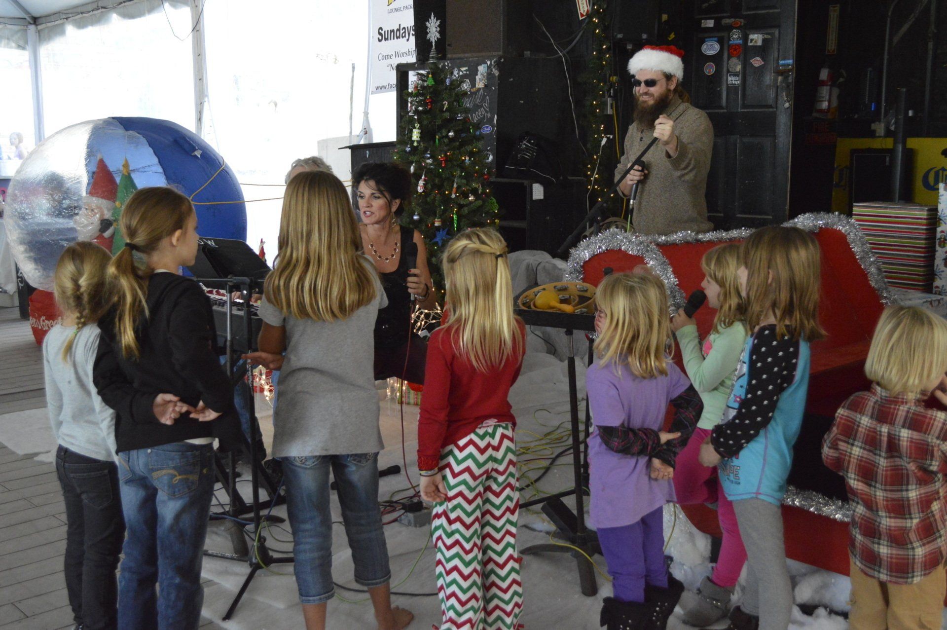 A group of children gathers around a woman singing and a man wearing a Santa hat near a Christmas tree and red sleigh.