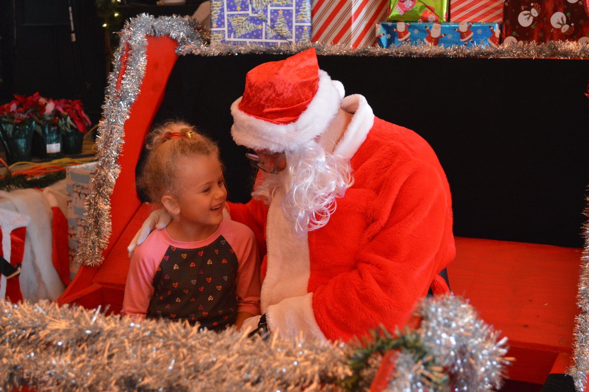 A child sits with Santa, both smiling, surrounded by Christmas decorations and gifts in a festive setting.