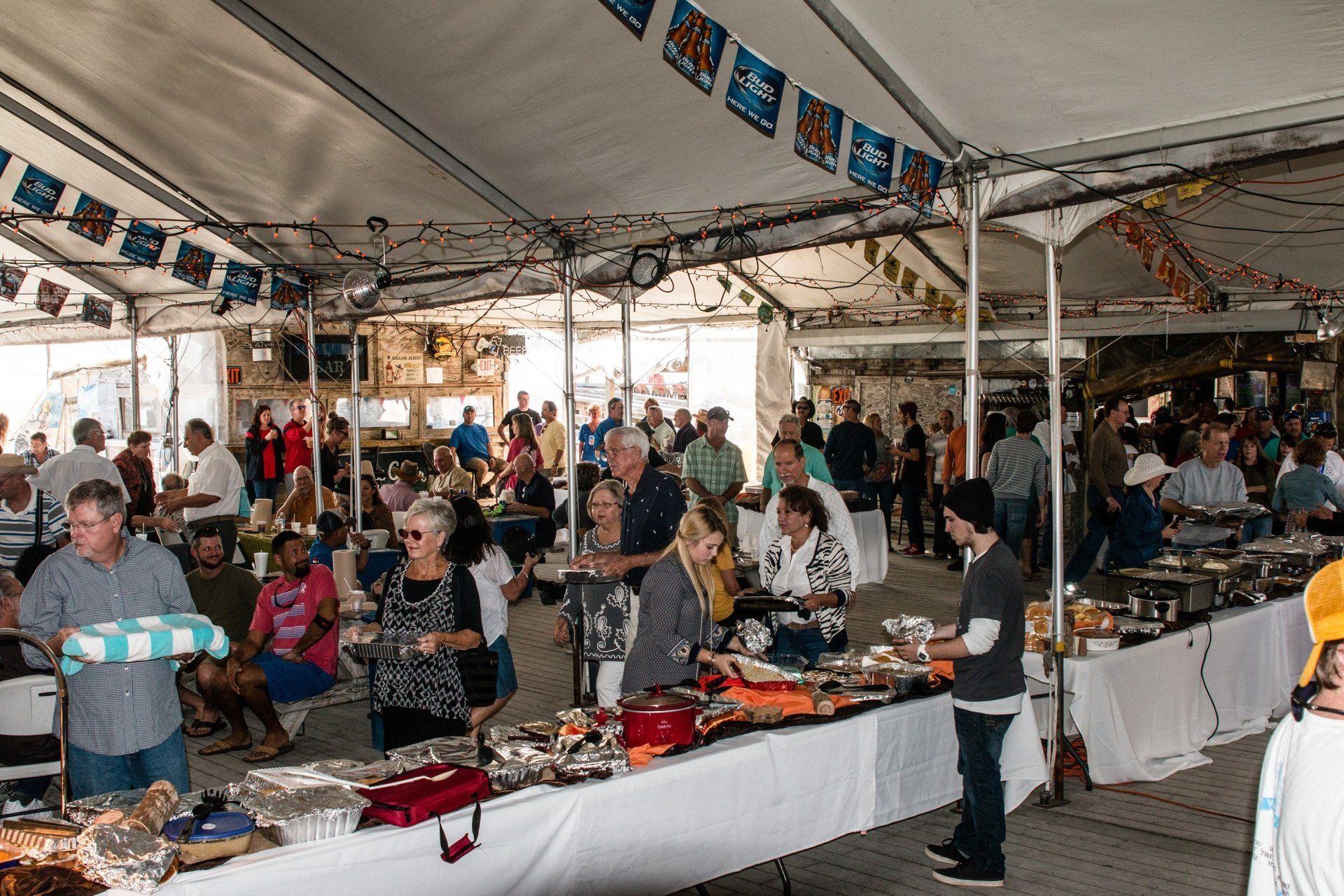 A large, crowded outdoor buffet under a tent, with people serving themselves food from long tables and chatting.