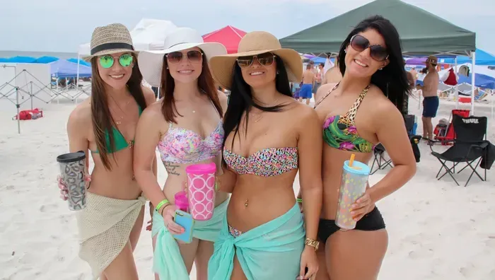Four people in swimwear and hats smile while standing on a sandy beach under a green tent canopy.