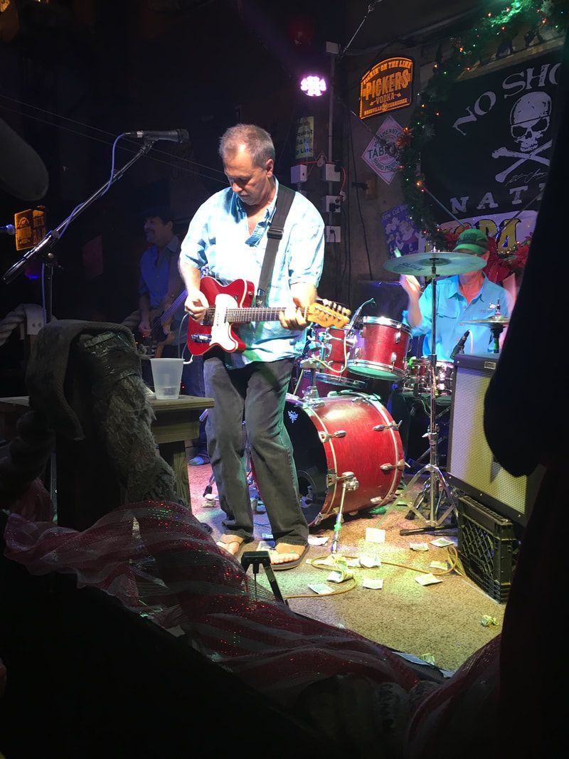 A guitarist plays a red electric guitar on a dimly lit stage next to a drummer, with a skull logo banner in the background.