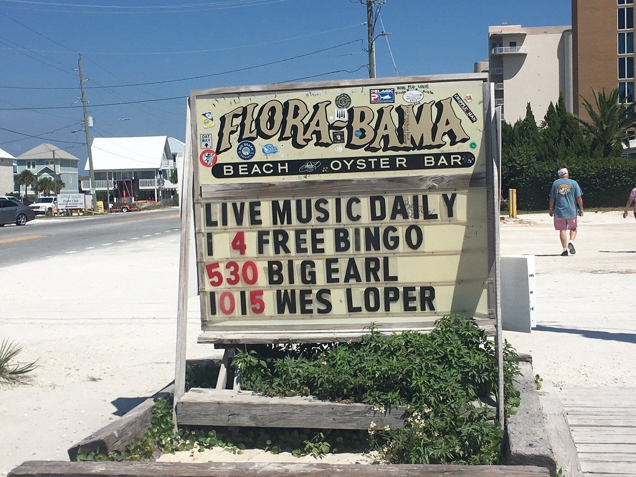 A Flora-Bama Beach Bar sign lists daily live music and events, with a person walking away in the sandy background.