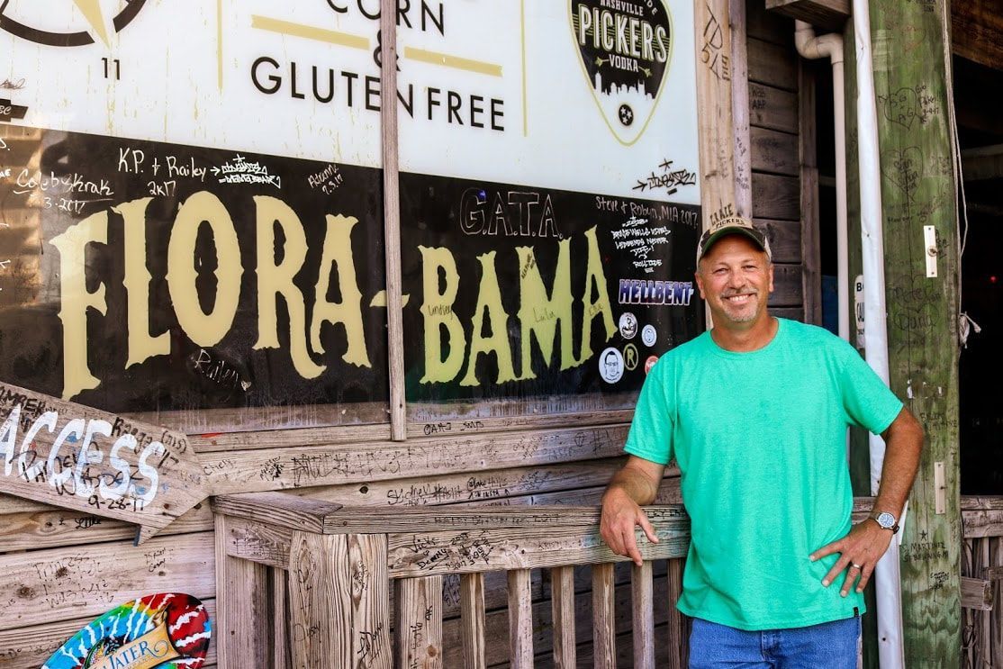 A smiling person in a green t-shirt stands outdoors in front of a sign that reads