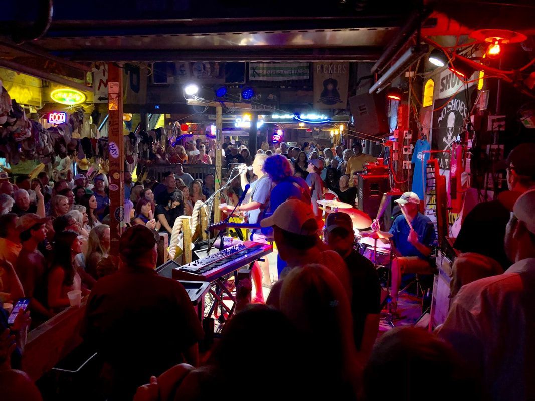A crowd watches a band perform on a small, dimly lit stage in a packed, rustic bar decorated with various signs.