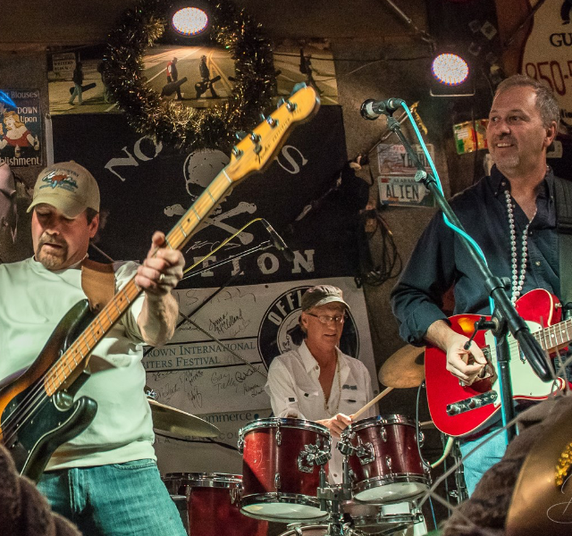 A three-piece band performing on a stage decorated with a skull logo, featuring a bassist, drummer, and guitarist.