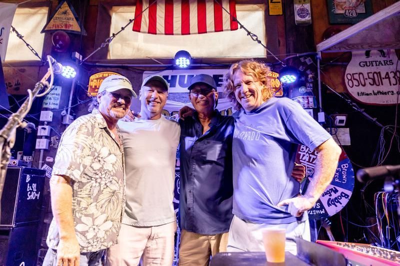 Four people stand together smiling under an American flag in a music venue with stage lights and equipment in the background.