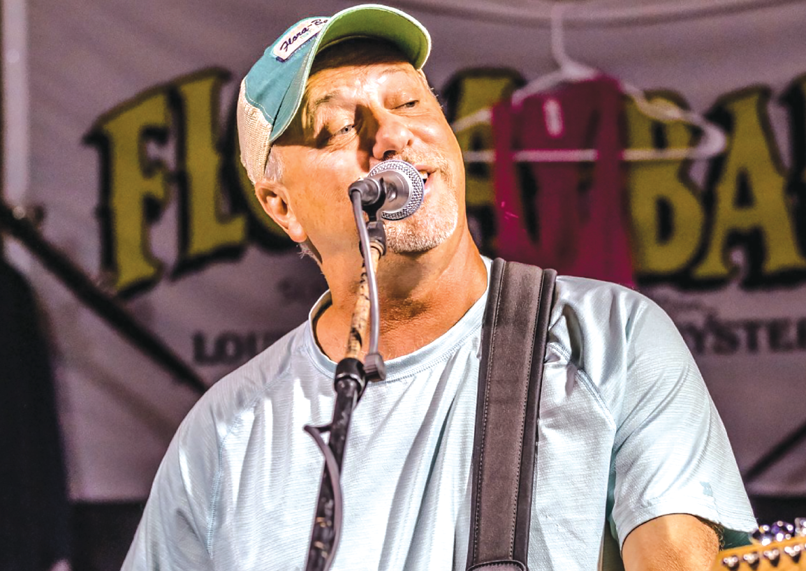 Big Earl A singer performs into a microphone on stage, wearing a light-colored t-shirt and a green baseball cap.