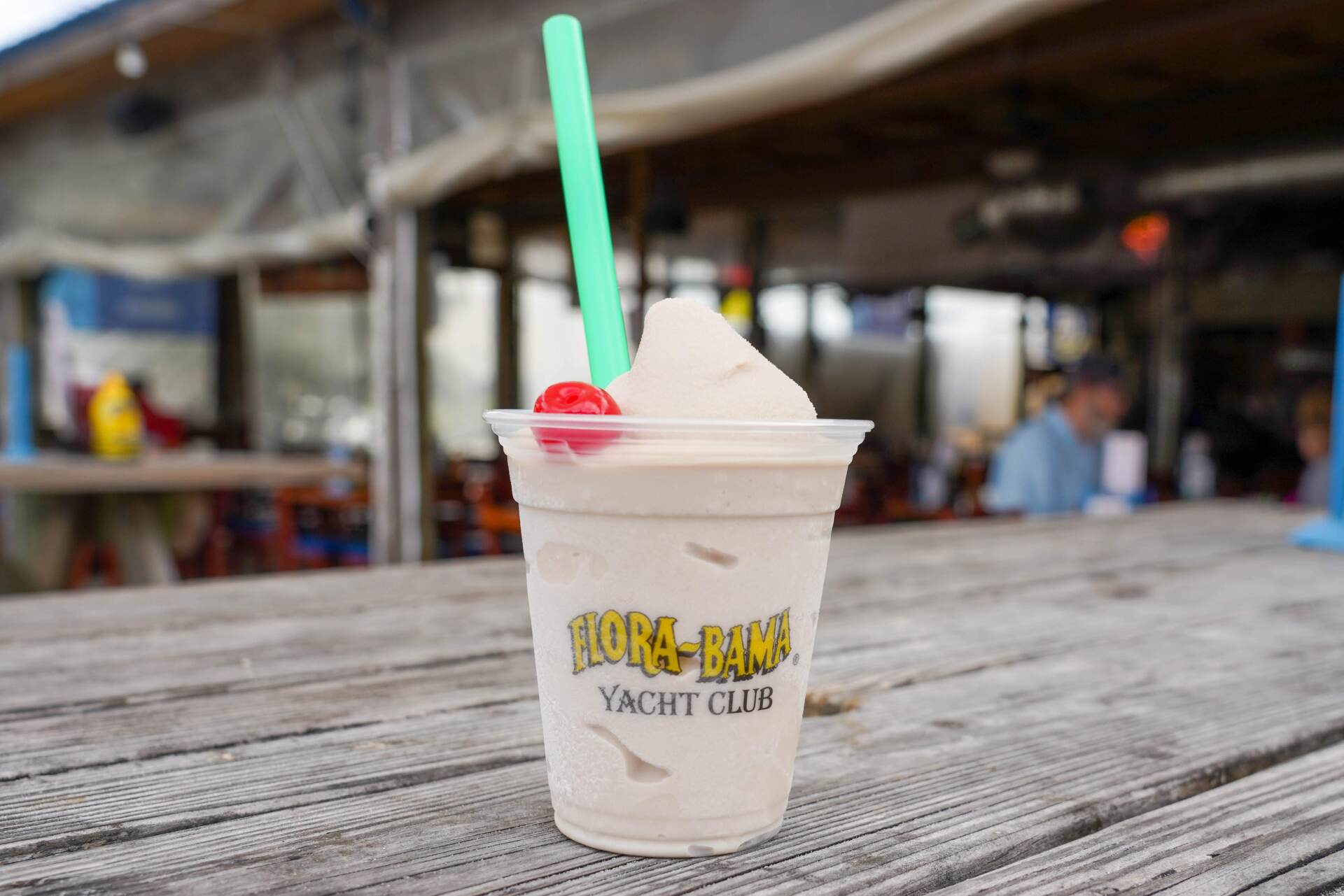 A frozen drink with a cherry and straw in a Flora-Bama Yacht Club plastic cup, sitting on a weathered outdoor wooden table.