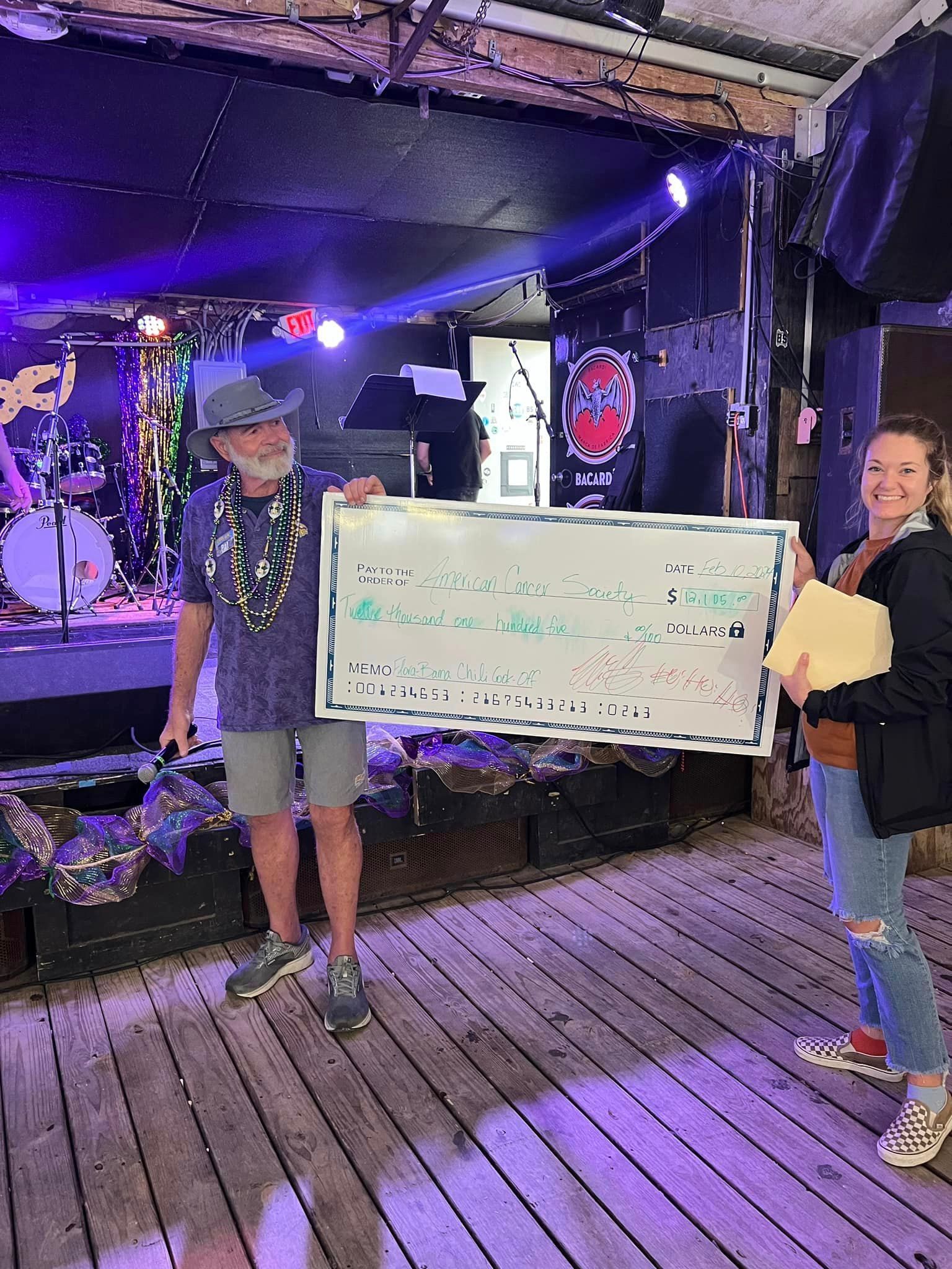 A person in a hat and a person in jeans hold a large ceremonial check on a wooden stage in front of band equipment.