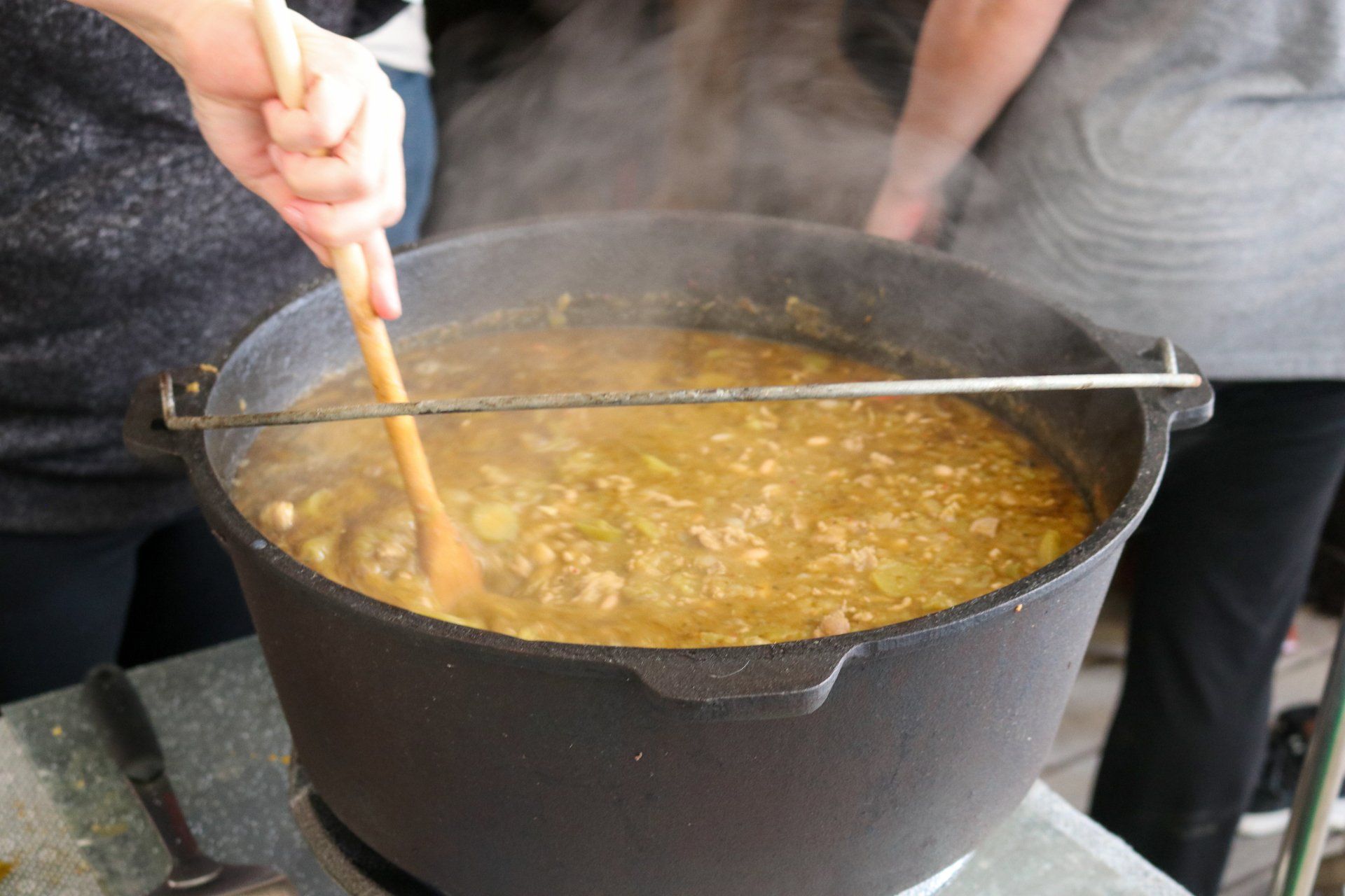 A person stirs a large, steaming pot of stew with a wooden spoon in an outdoor setting.