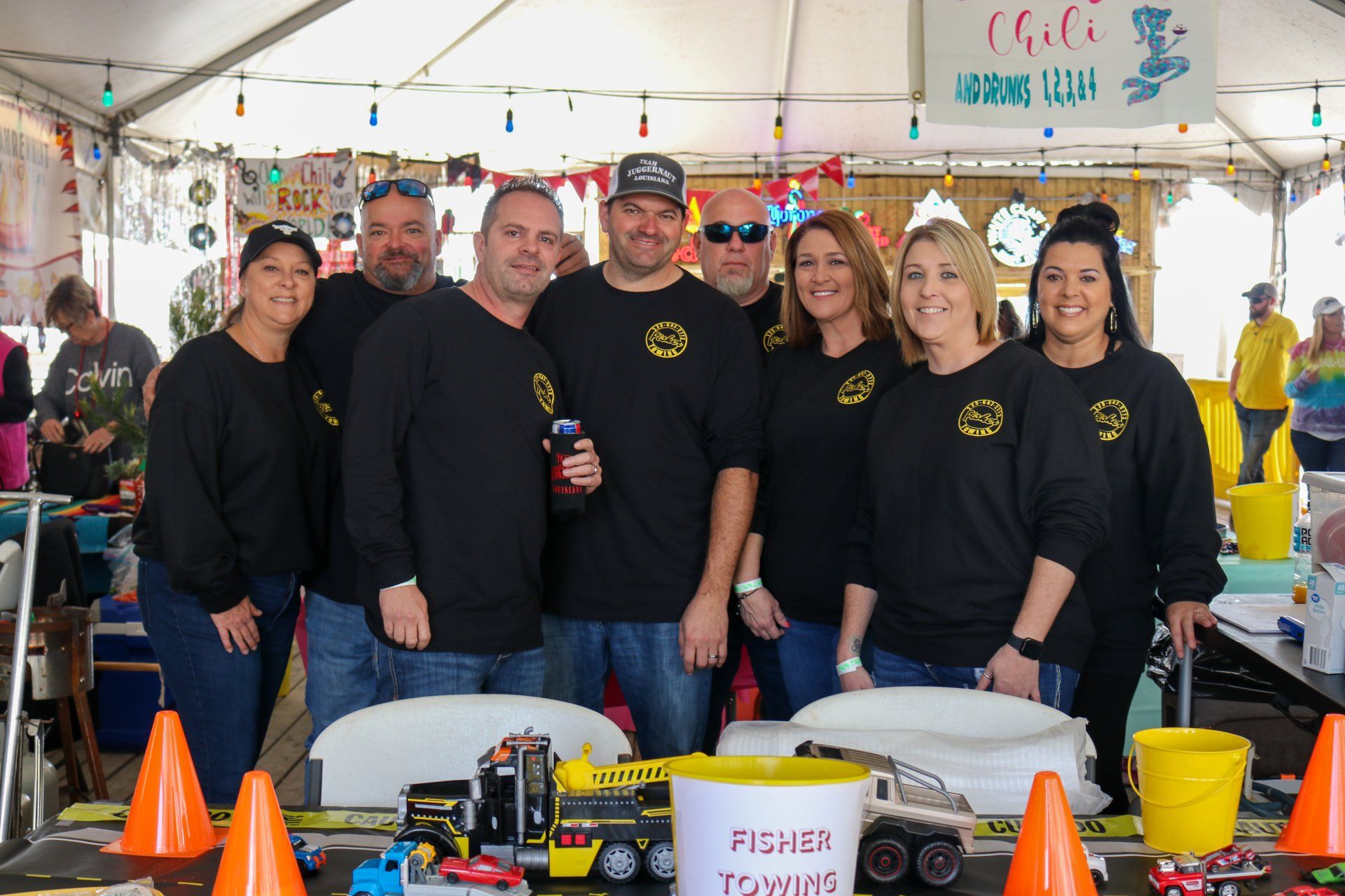 A group of people in black shirts stands under a tent for a chili cook-off, with toy trucks on a table in the foreground.