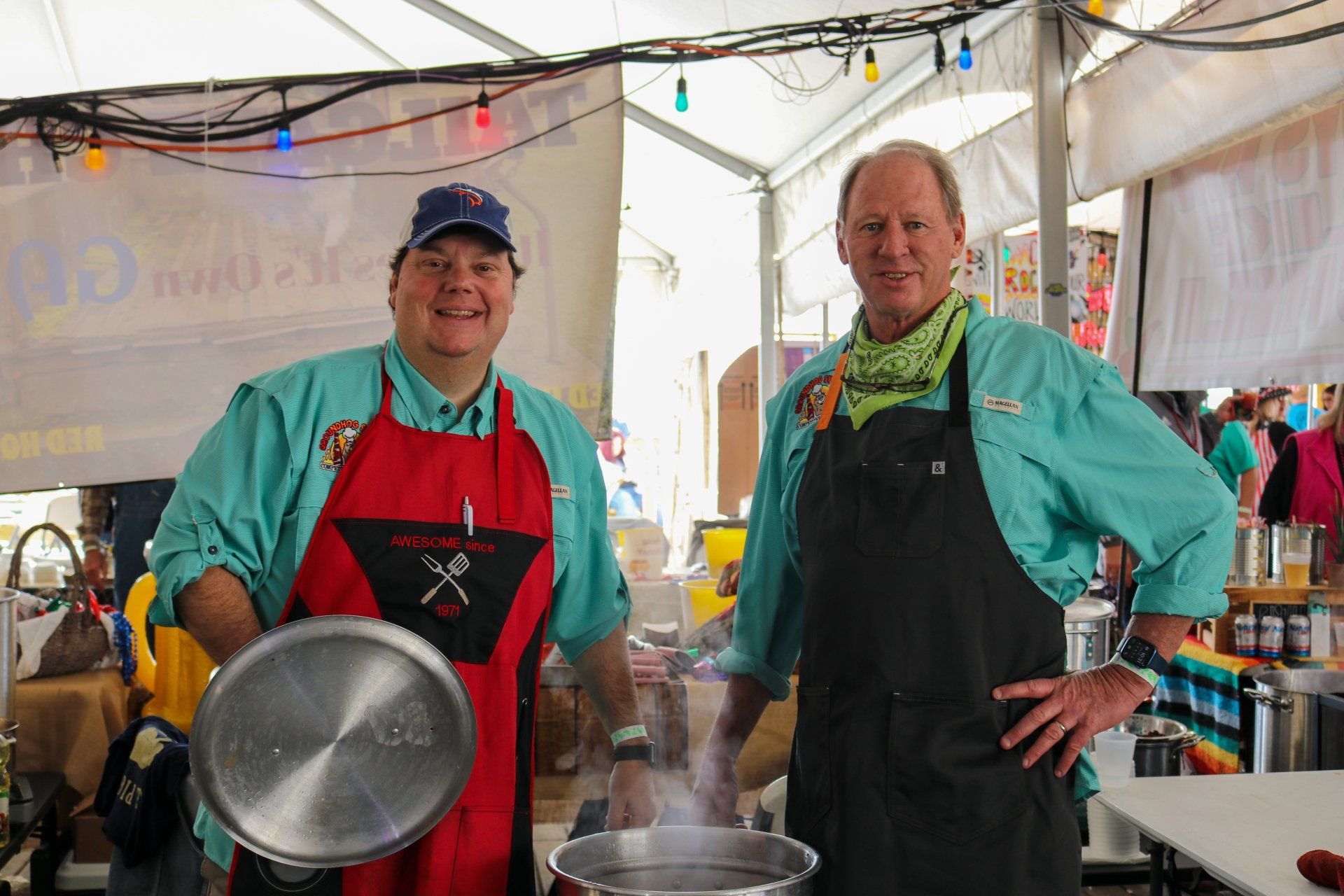 Two people in matching teal shirts and aprons stand under a tent, one holding a pot lid over a steaming pot.