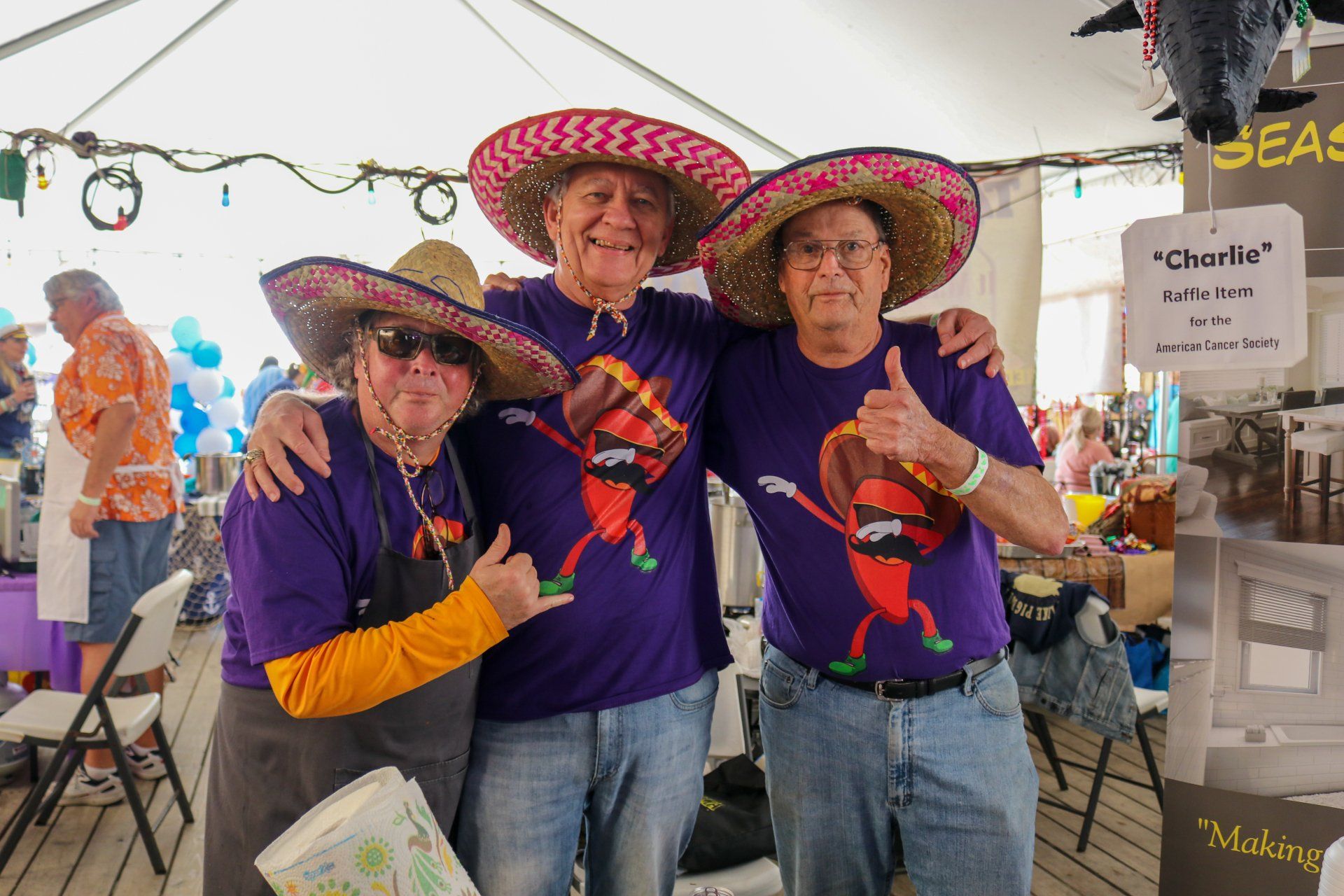 Three people in matching purple shirts and colorful sombreros pose together under a tent, one giving a thumbs up.