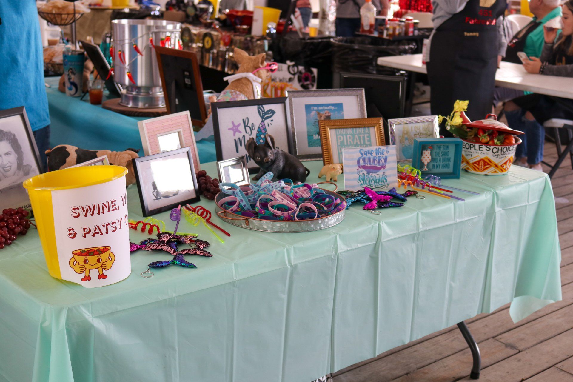 A light blue table at an outdoor event holds party decorations, framed signs, a yellow bucket, and small giveaways.