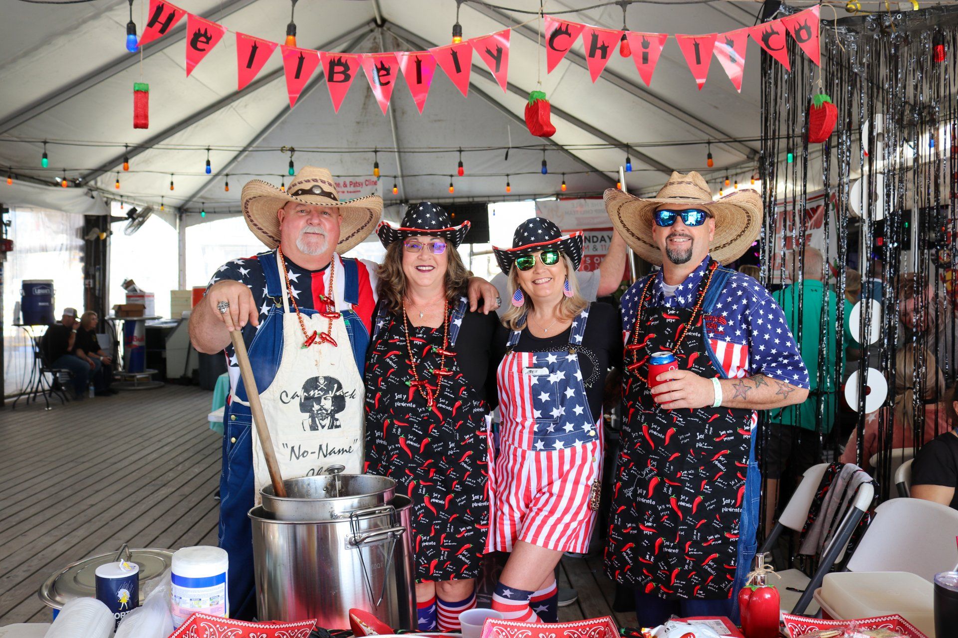 Four people in patriotic aprons and cowboy hats prepare chili at an outdoor stand under a banner.