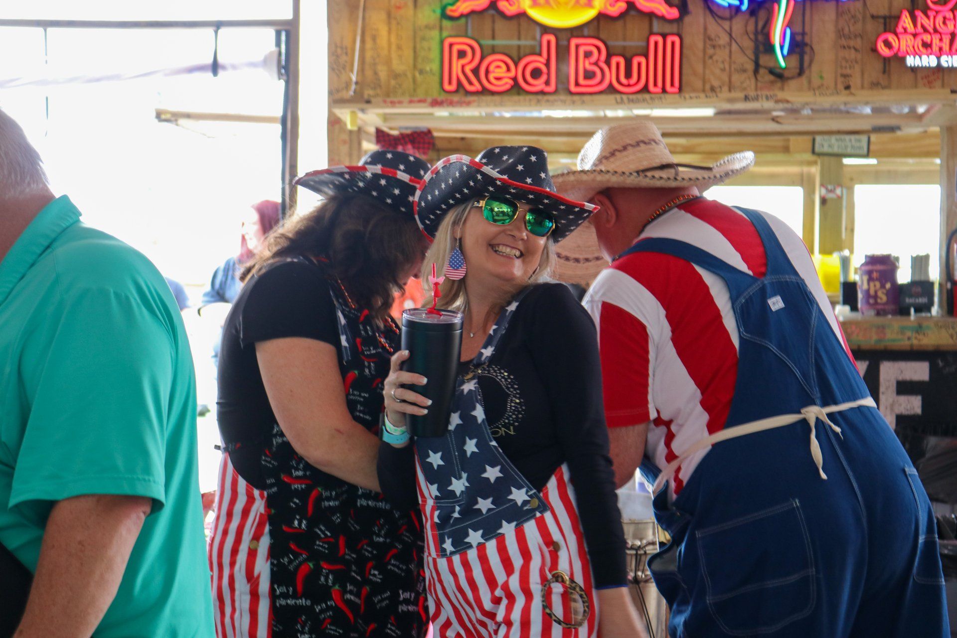 People in patriotic American-flag-themed overalls and hats socializing in a brightly lit bar with a Red Bull sign.