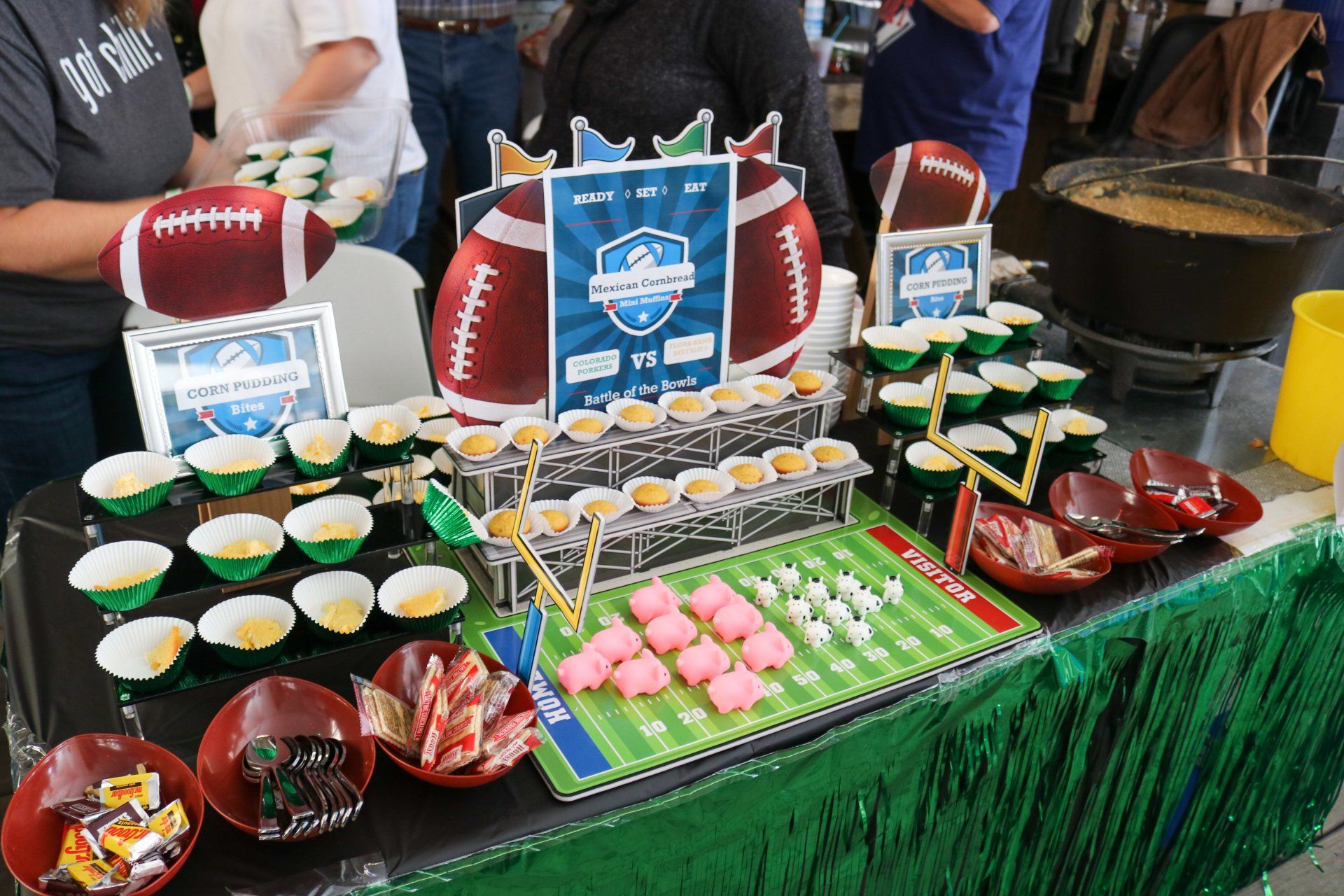 A game-day themed table featuring football decorations, snacks in green paper cups, small candies, and a football field mat.