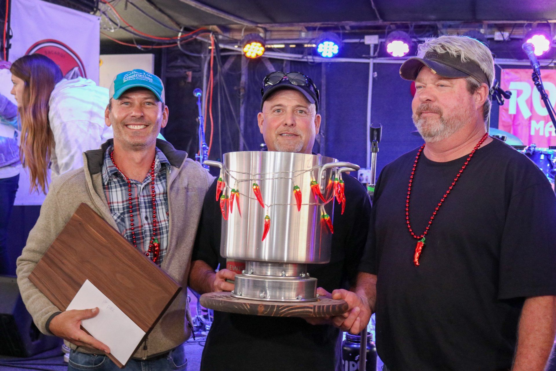 Three people stand on a stage, with the center person holding a large chili-decorated trophy, celebrating a contest win.