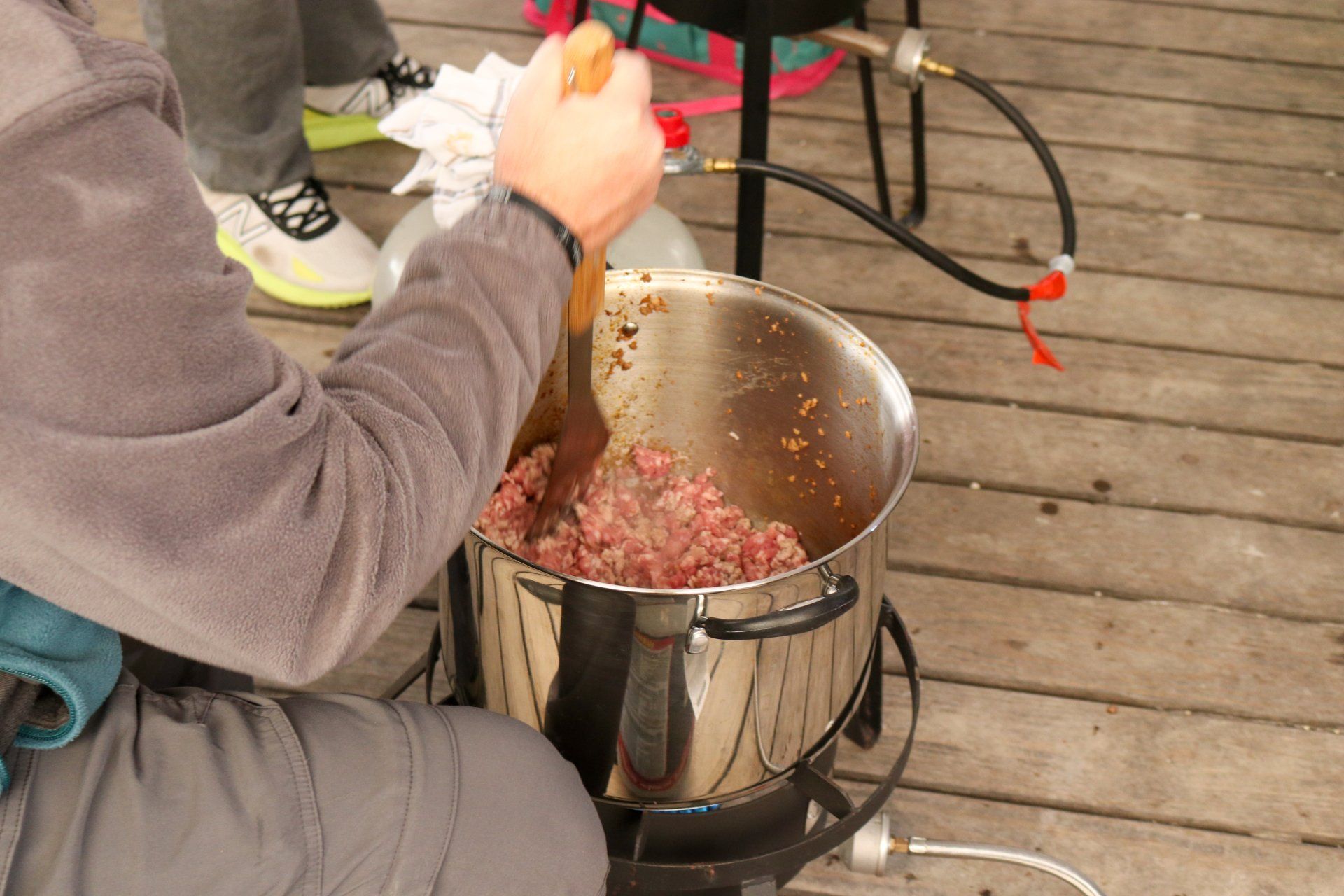 A person stirs ground meat in a large stainless steel pot on a propane burner outdoors on a wooden deck.