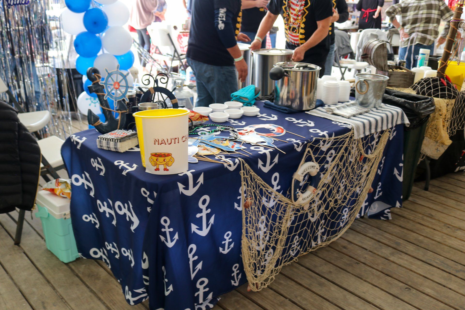 A table decorated with a blue anchor-print tablecloth and nautical netting, featuring food preparation items at a fair.
