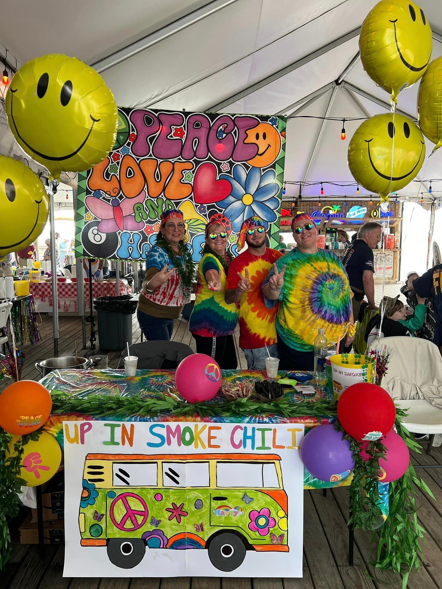 Four people in tie-dye shirts stand behind a table decorated with balloons and a sign reading 