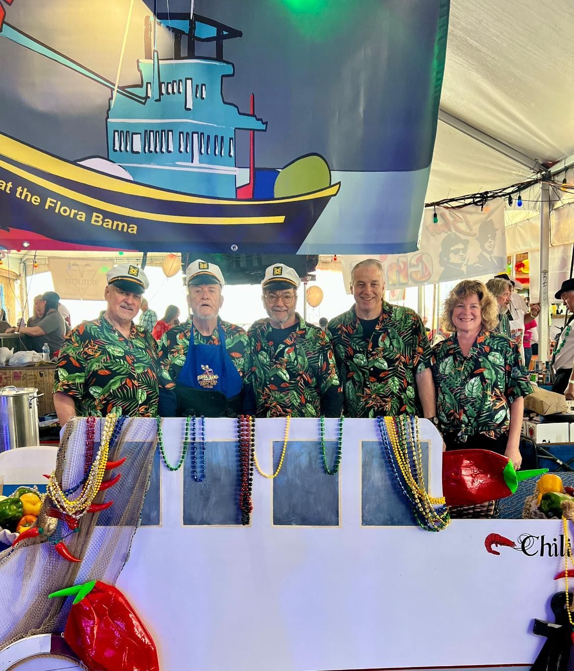 Five people in matching tropical shirts and hats pose behind a decorated boat cutout under a tent at a festival.