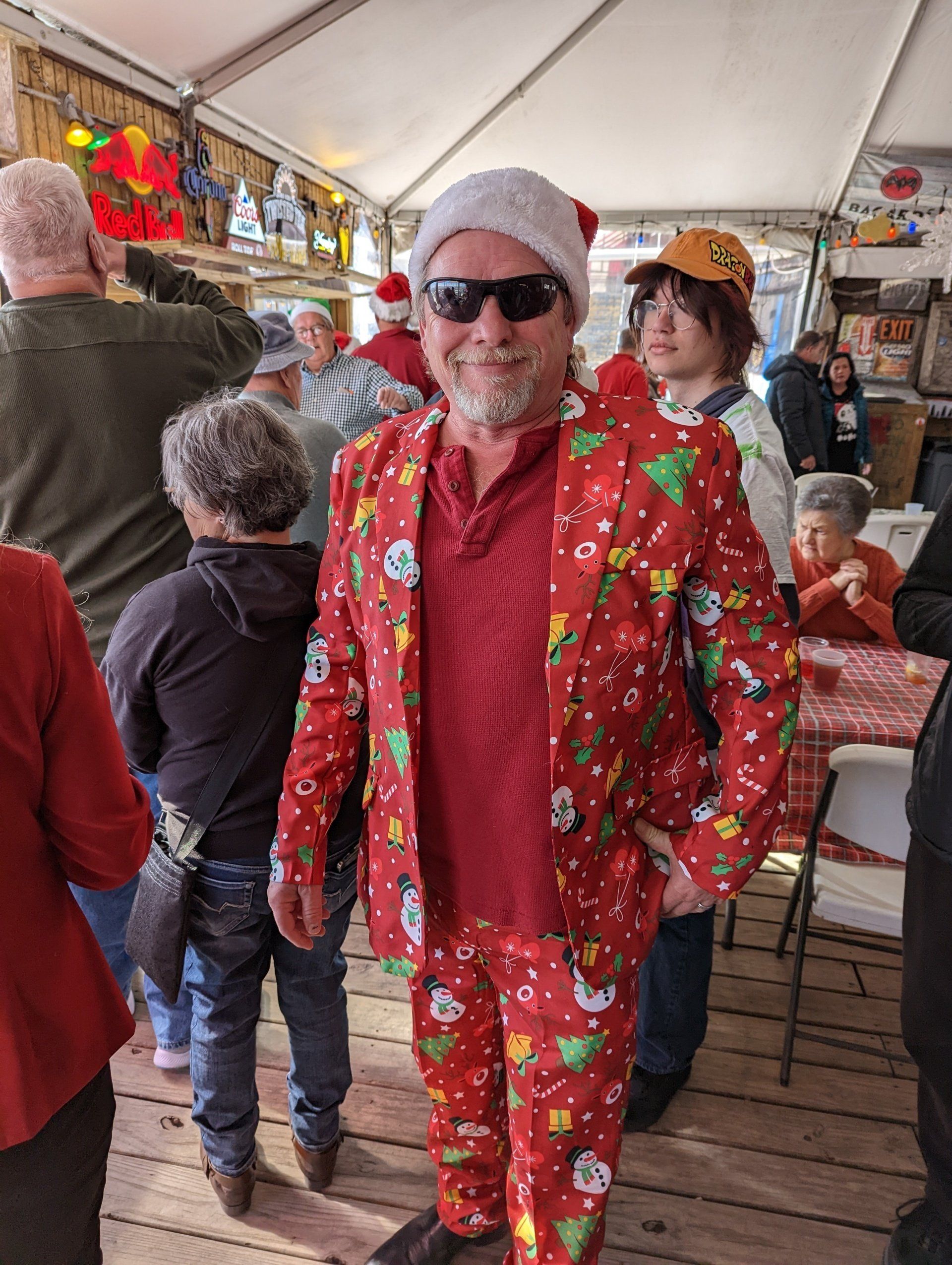 A person wearing a Santa hat and a red holiday-themed suit stands inside a tent with others at a festive gathering.