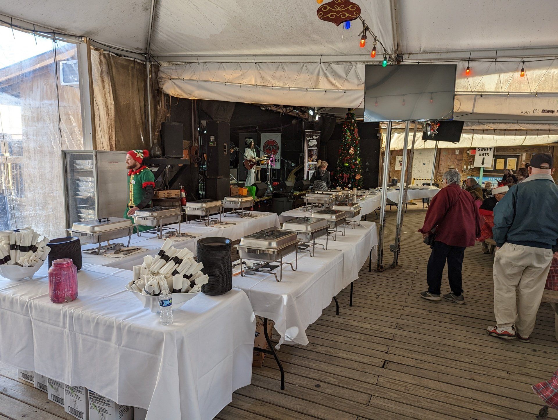 A catering buffet set up under a tent with white tablecloths, stainless steel food warmers, and patrons standing nearby.