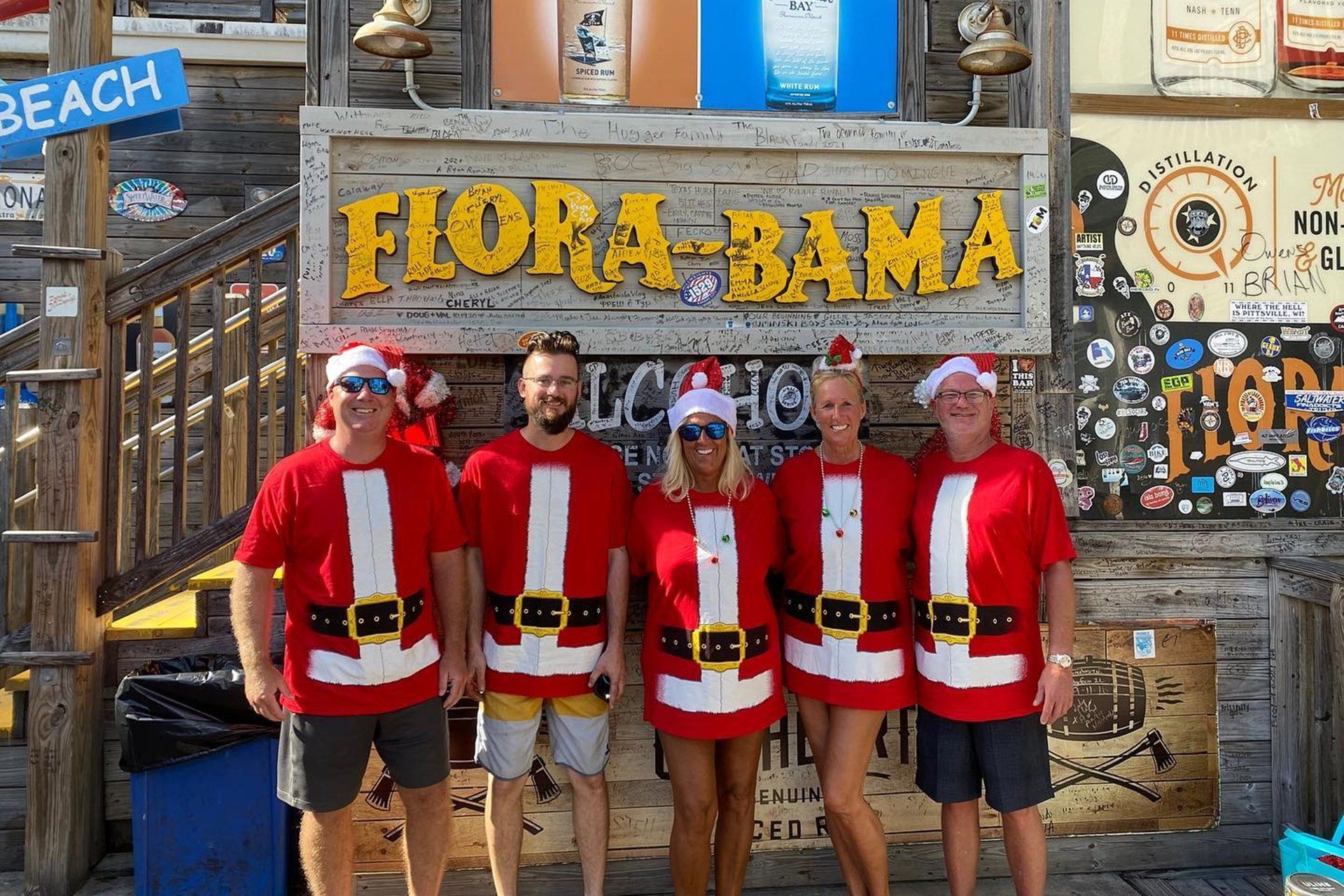 Five people in Santa-themed shirts stand together smiling in front of the Flora-Bama sign at a beach location.