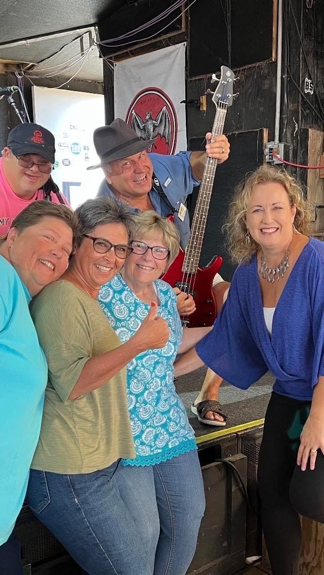 Six people pose for a group photo indoors; one holds a guitar, and everyone is smiling at the camera.