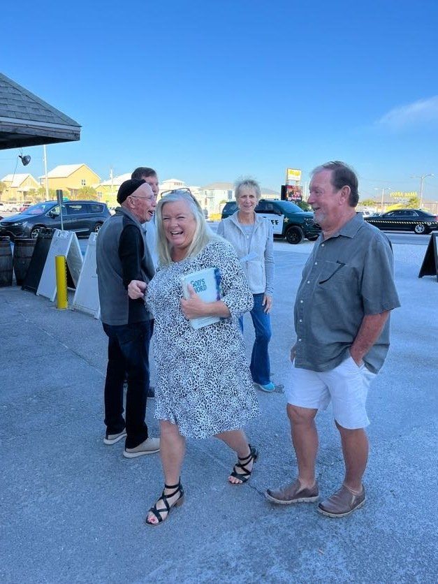 A group of four adults laughing and talking in a sunny, paved parking lot.